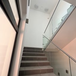 AD-REM FIRMA USŁUGOWA ADAM GRACZYK - Nowoczesna klatka schodowa z ciemnymi, prostokątnymi płytkami na stopniach i szklaną balustradą z metalową poręczą. Białe ściany i duże okno.