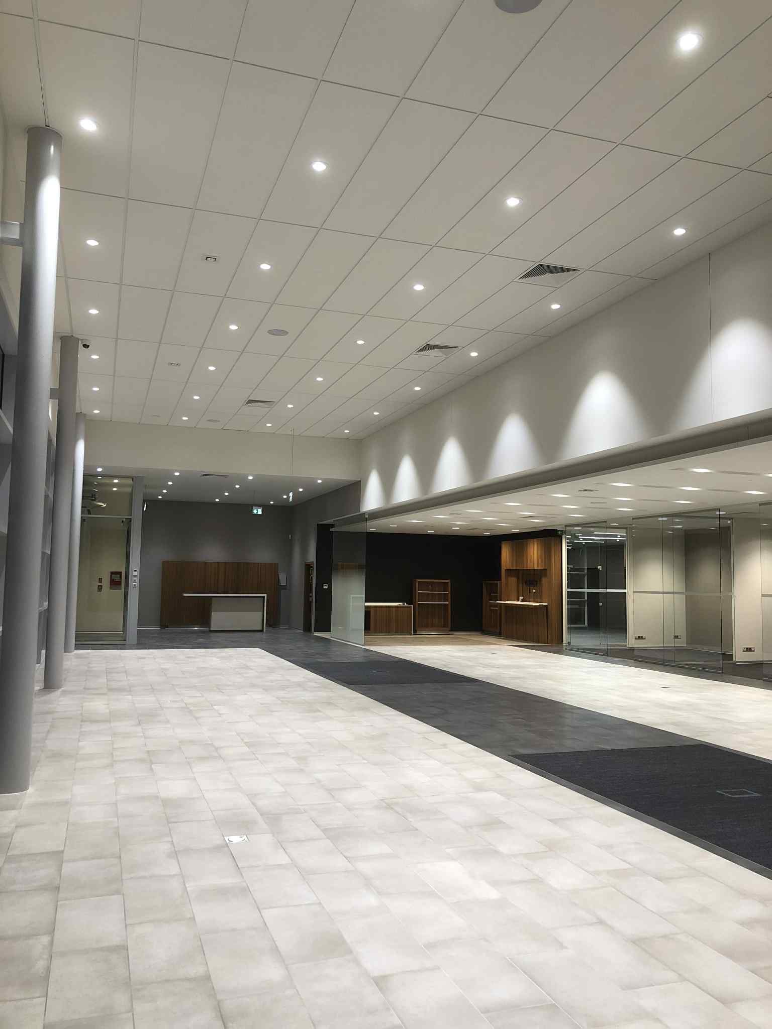 Jasne, przestronne wnętrze biurowe z płytkami podłogowymi, szklanymi ścianami i nowoczesnym oświetleniem sufitowym. Widoczne elementy wykończenia i recepcja.