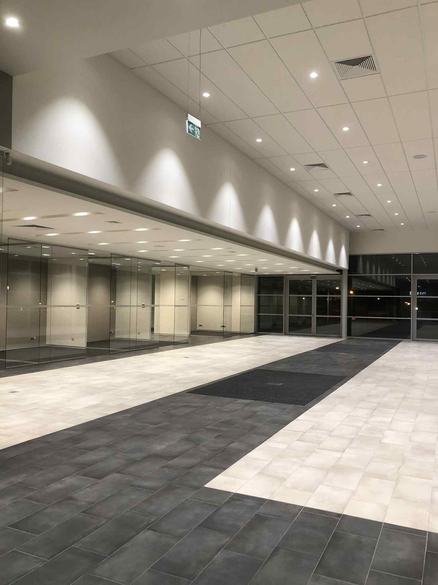 Jasne, przestronne wnętrze biurowe z przeszklonymi ścianami, nowoczesnym oświetleniem i podłogą z płytek w odcieniach szarości i bieli. Widoczny znak ewakuacyjny.
