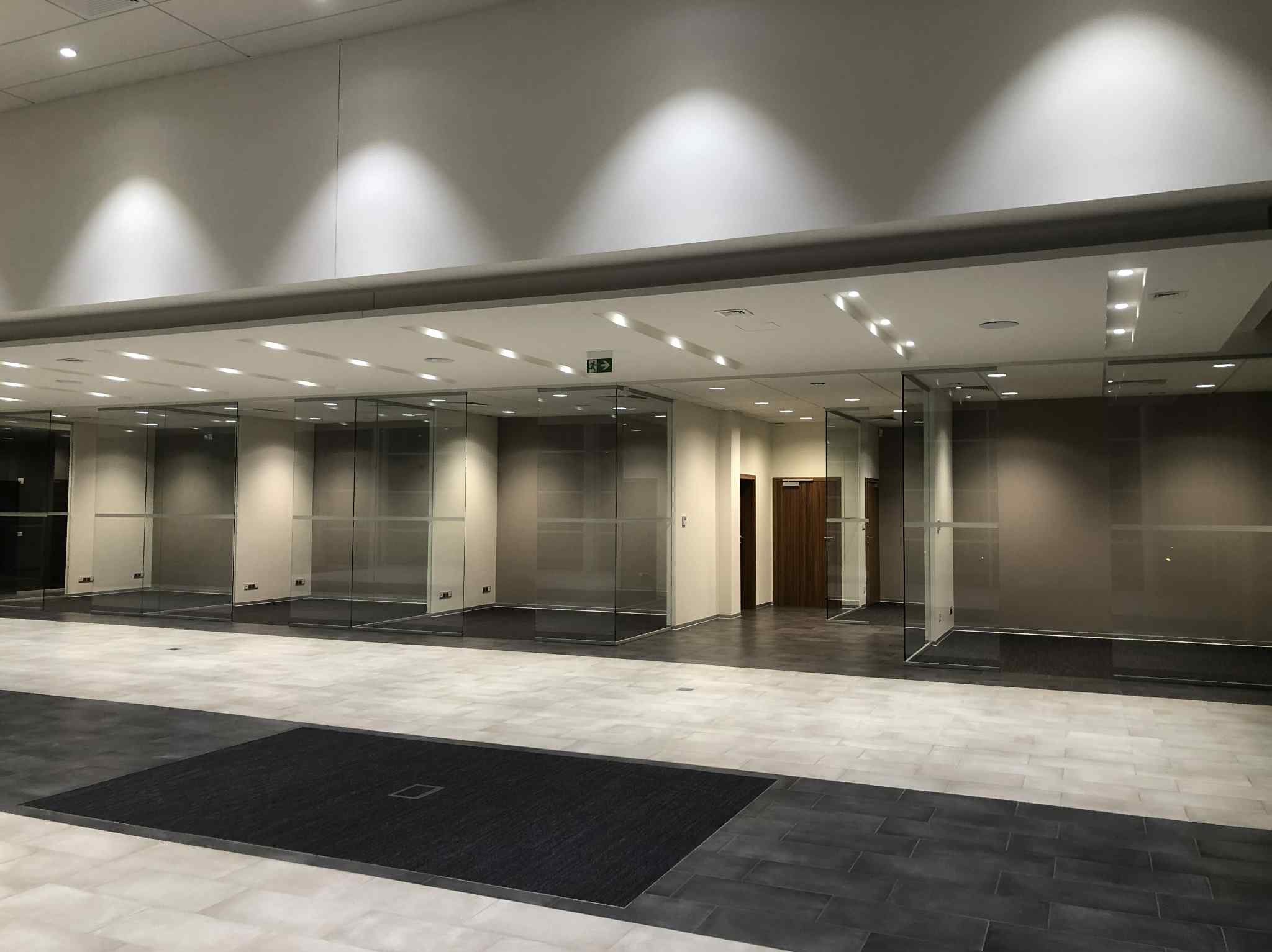 Nowoczesne wykończenie wnętrza biurowego z przeszklonymi ściankami działowymi, jasną podłogą i oświetleniem sufitowym.