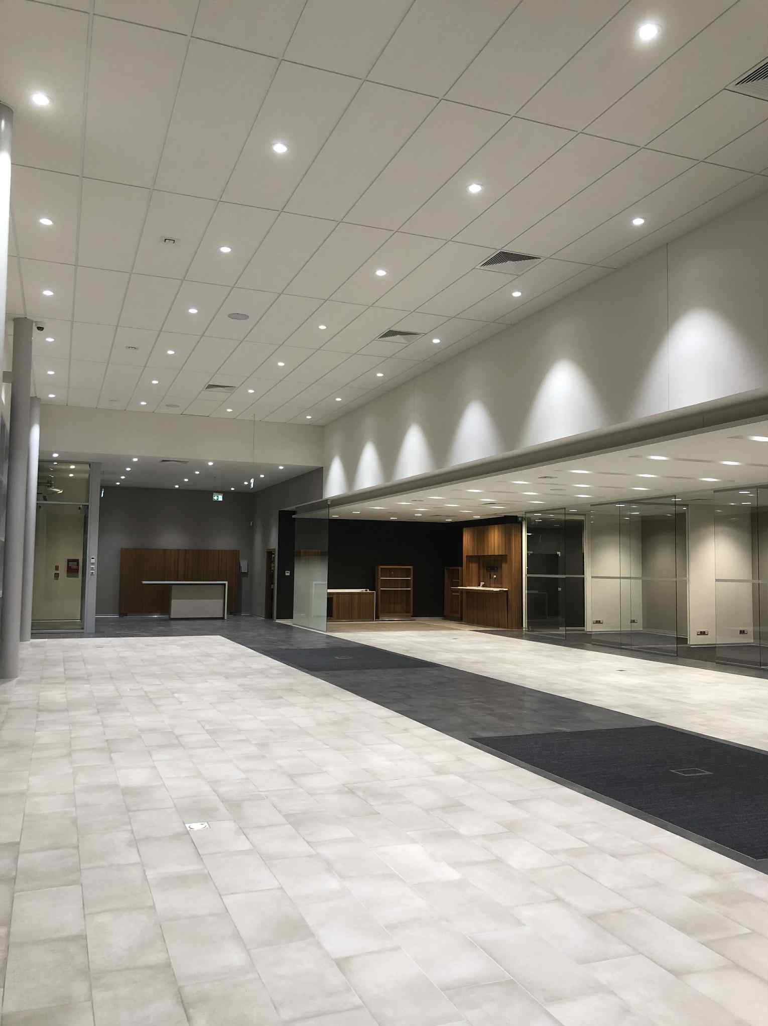 Przestronne, jasne wnętrze biurowe z podwieszanym sufitem, oświetleniem punktowym i szarą podłogą z płytek. Widoczne przeszklone ściany i elementy wykończenia z drewna.