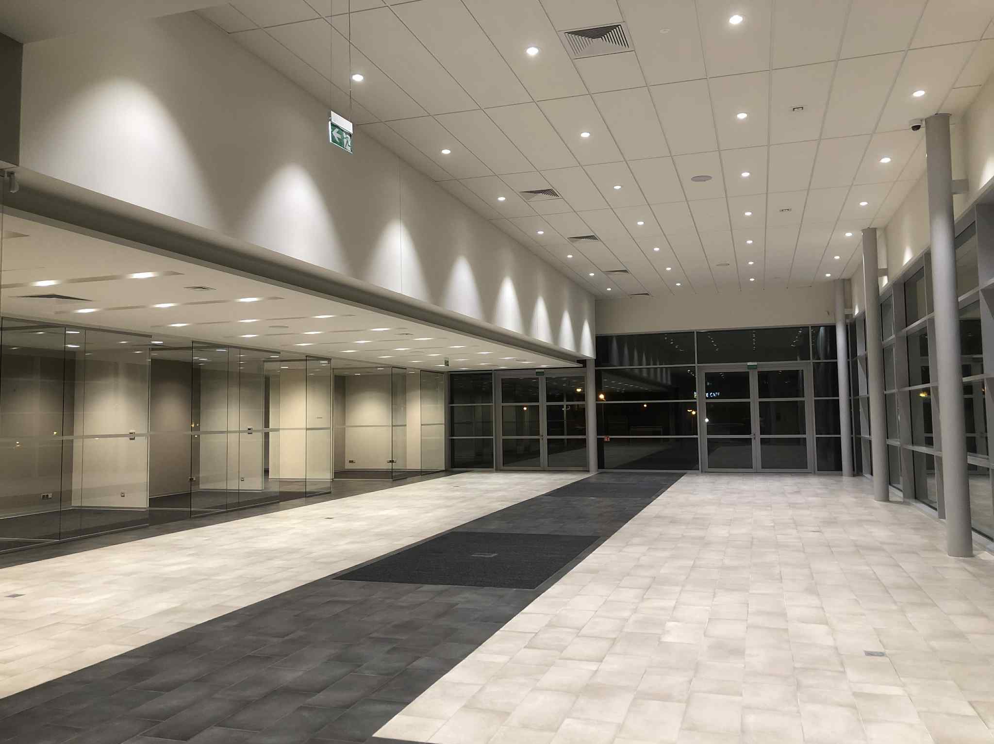 Jasne, przestronne wnętrze biurowe z przeszklonymi ścianami i nowoczesnym oświetleniem sufitowym. Podłoga wyłożona płytkami w odcieniach szarości i beżu.