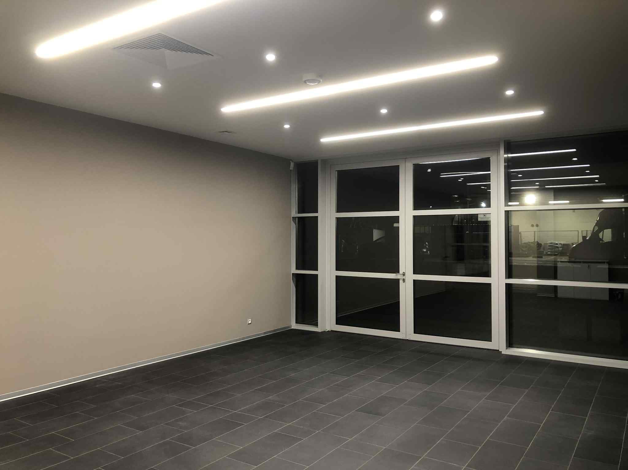 Wykończone wnętrze biurowe z szarą ścianą, ciemną podłogą z płytek i przeszklonymi drzwiami. Oświetlenie liniowe LED na suficie. Minimalistyczny design.