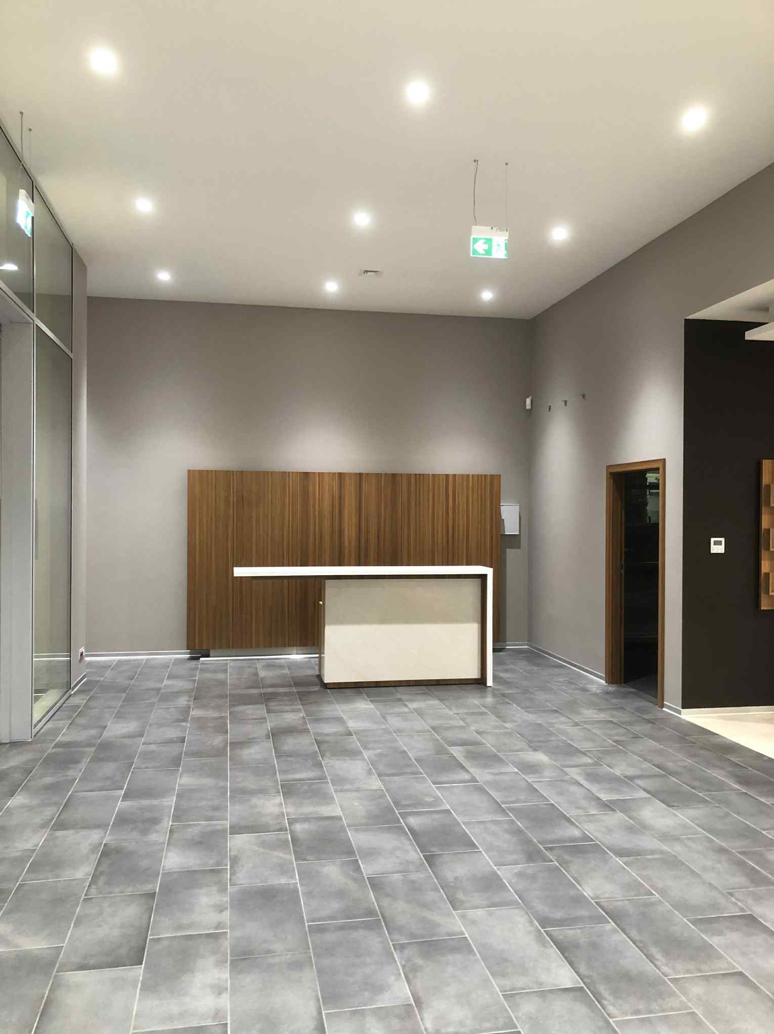 Wykończone wnętrze biura z szarymi płytkami podłogowymi, drewnianą ścianą i recepcją. Widoczny znak ewakuacyjny. Stonowana kolorystyka, nowoczesny design.