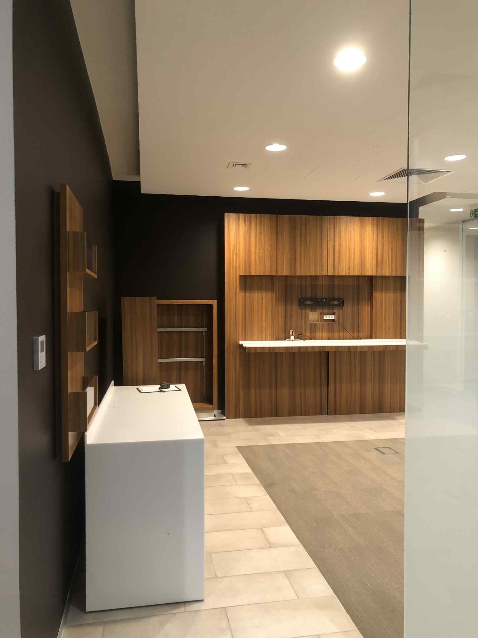 Nowoczesne wykończenie wnętrza biura: ciemna ściana z drewnianymi półkami, biała lada recepcyjna i drewniana zabudowa ściany z ukrytymi szafkami.