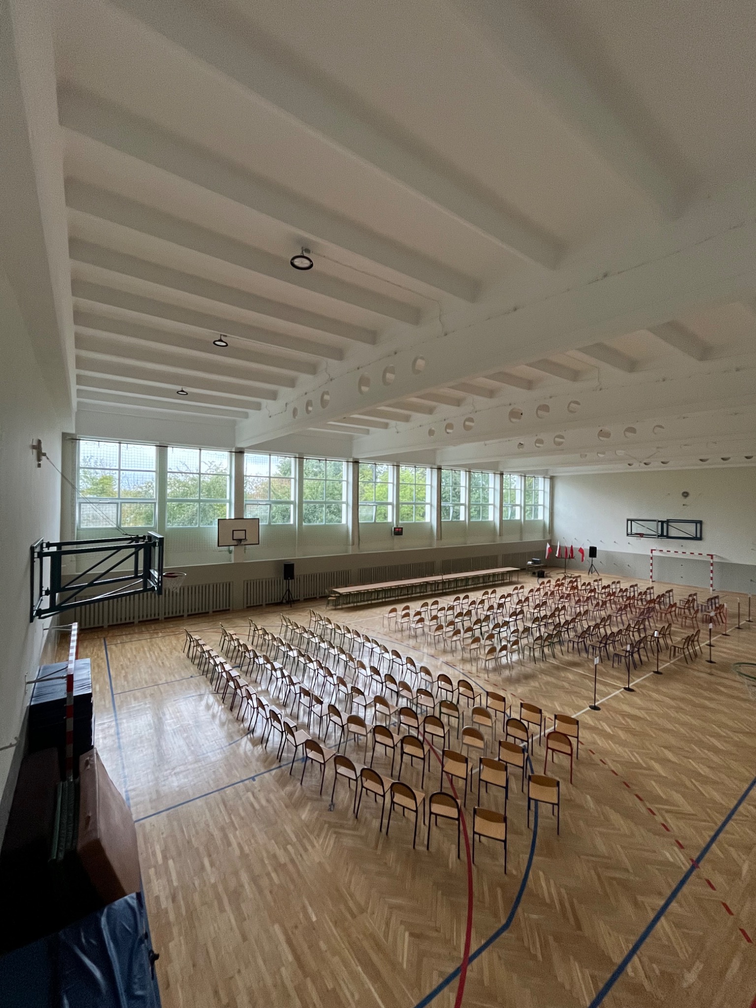 Sala gimnastyczna z parkietem, przygotowana na uroczystość: rzędy krzeseł, stół, flaga. Widok z góry, oświetlenie naturalne z okien.