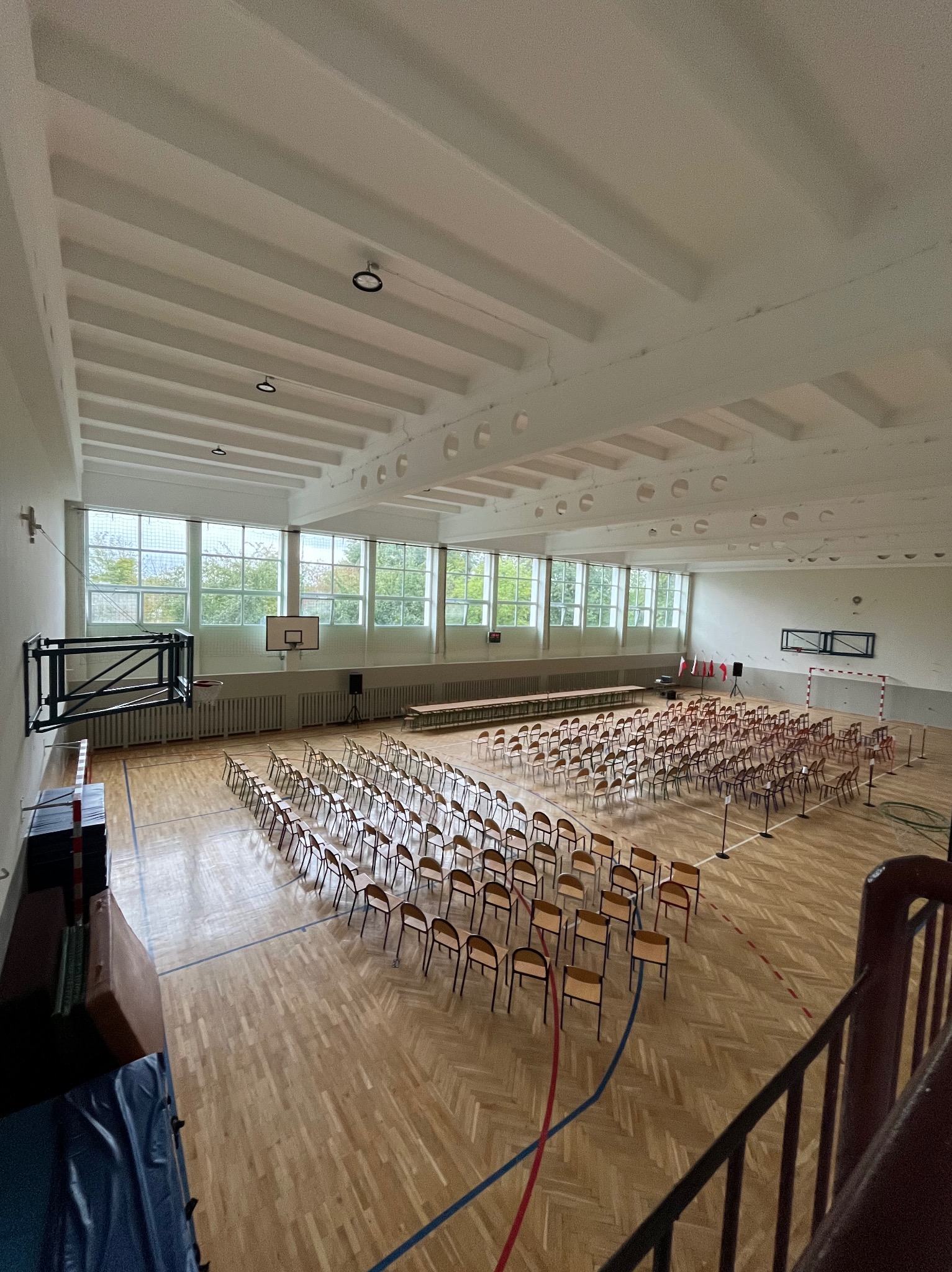 Sala gimnastyczna z parkietem, rzędy krzeseł ustawione do wydarzenia, widok z góry. Białe ściany i sufit z oknami w tle. Kosz do koszykówki i bramka.