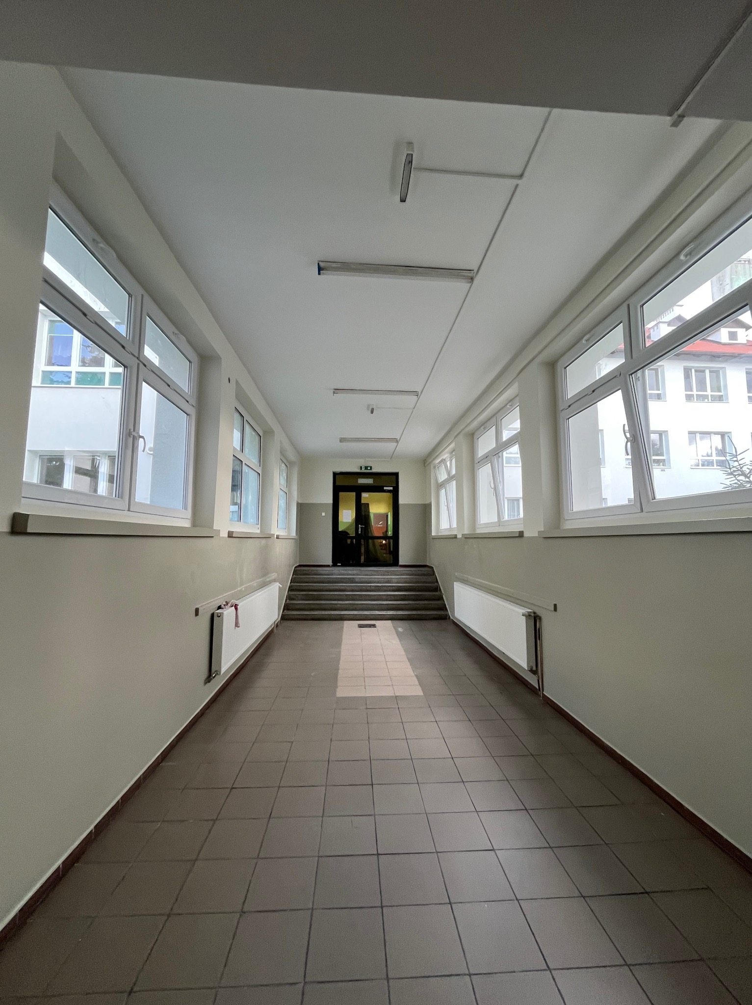 Długi korytarz z oknami po bokach, prowadzący do schodów i drzwi. Ściany pomalowane na jasny kolor, podłoga wyłożona brązowymi płytkami. Widoczne grzejniki pod oknami.
