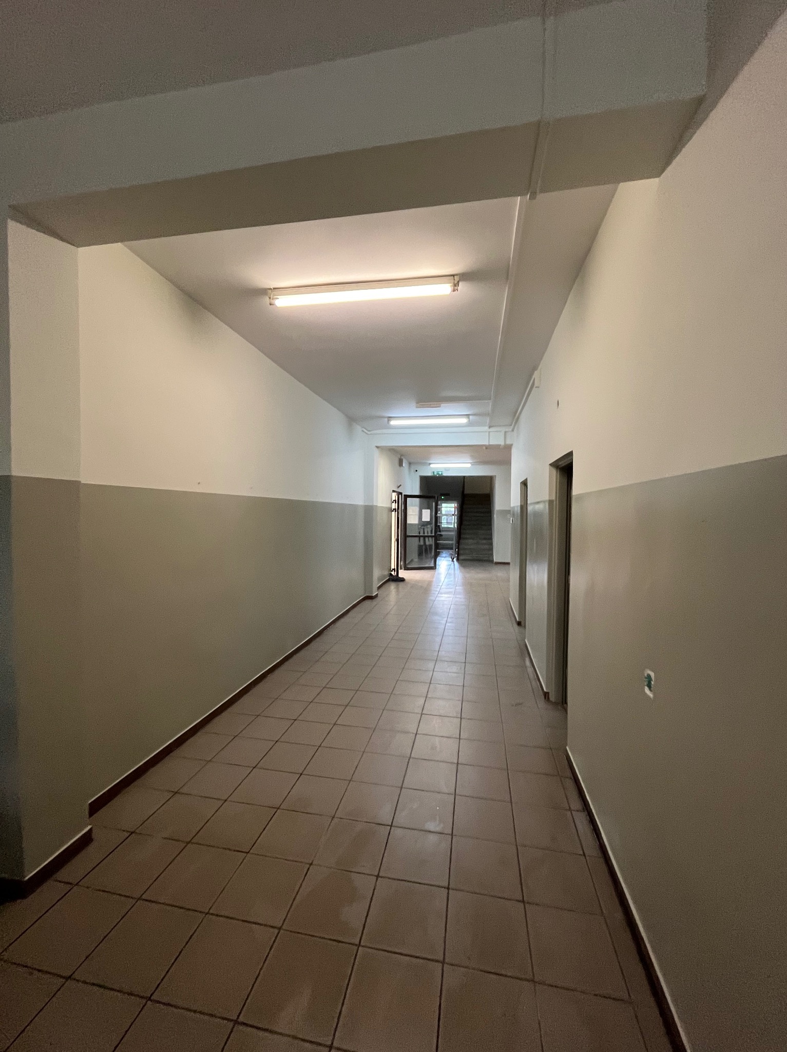 Korytarz po odświeżeniu w Lniano: ściany pomalowane na dwa kolory, jasna góra i ciemniejszy dół. Brązowa podłoga z płytek, sufit z lampami. Widok w głąb korytarza.