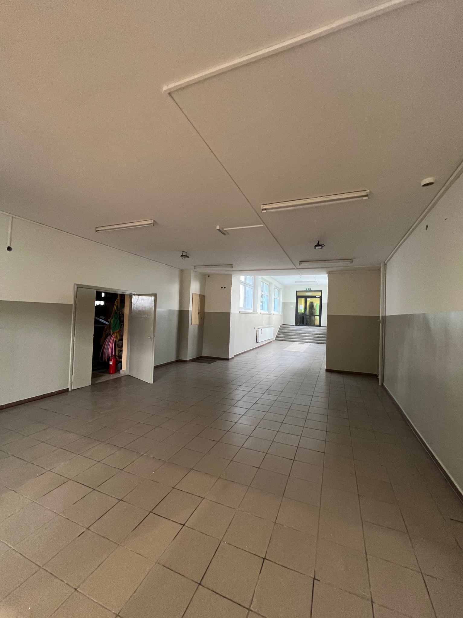 Przestronne wnętrze z kafelkowaną podłogą i dwukolorowymi ścianami. Widoczne otwarte drzwi do pomieszczenia gospodarczego i dalsza część korytarza z oknami.