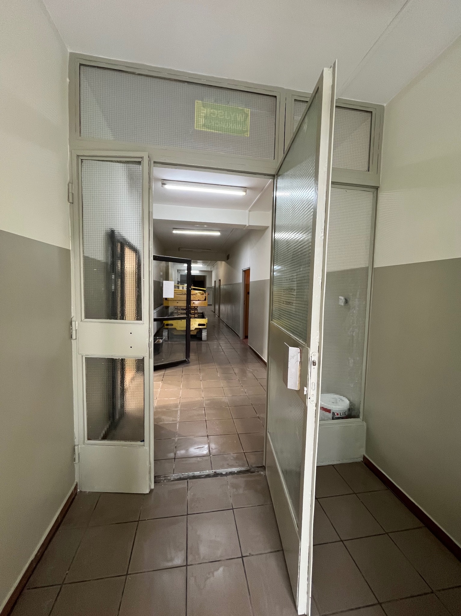 Korytarz w budynku z otwartymi, przeszklonymi drzwiami. Widoczny podnośnik nożycowy w oddali. Ściany pomalowane do połowy na szaro, reszta na biało.