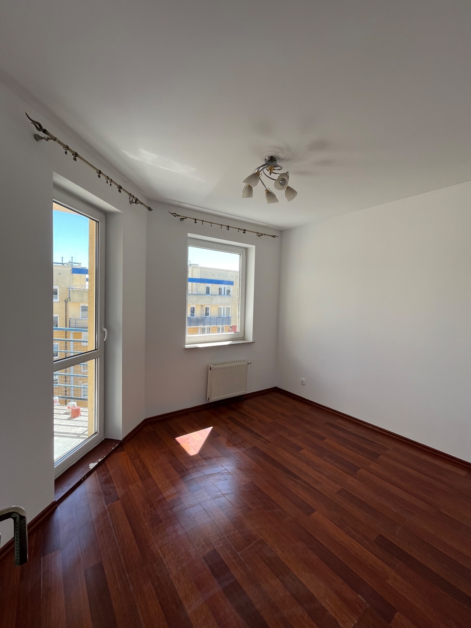 Pusty pokój po remoncie: świeżo pomalowane białe ściany, błyszcząca, ciemna podłoga z paneli, okna z widokiem na budynek. Minimalistyczna lampa sufitowa.