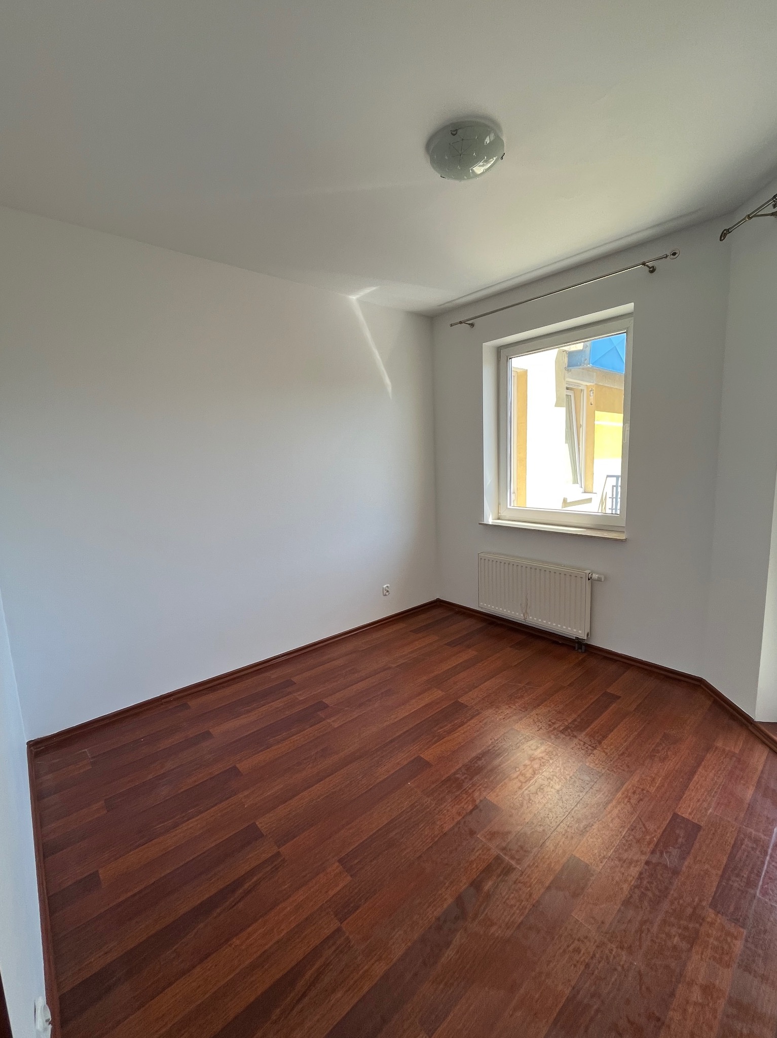 Puste, jasne wnętrze po remoncie: ciemne panele podłogowe, białe ściany, okno z grzejnikiem. Widoczny fragment karnisza i lampa sufitowa.