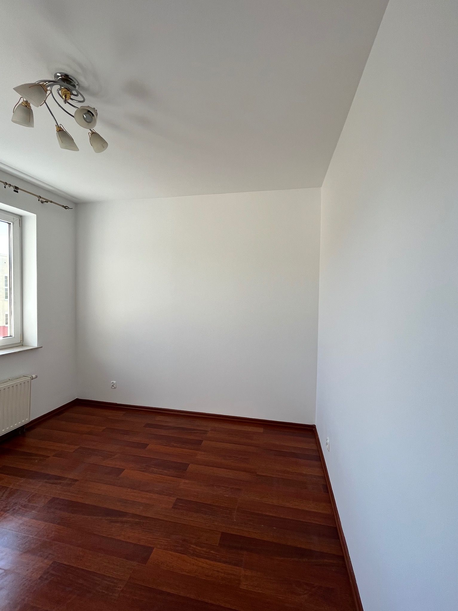 Pusty pokój po remoncie: świeżo pomalowane białe ściany, ciemna drewniana podłoga, okno z grzejnikiem i lampa sufitowa. Widok na narożnik pokoju.