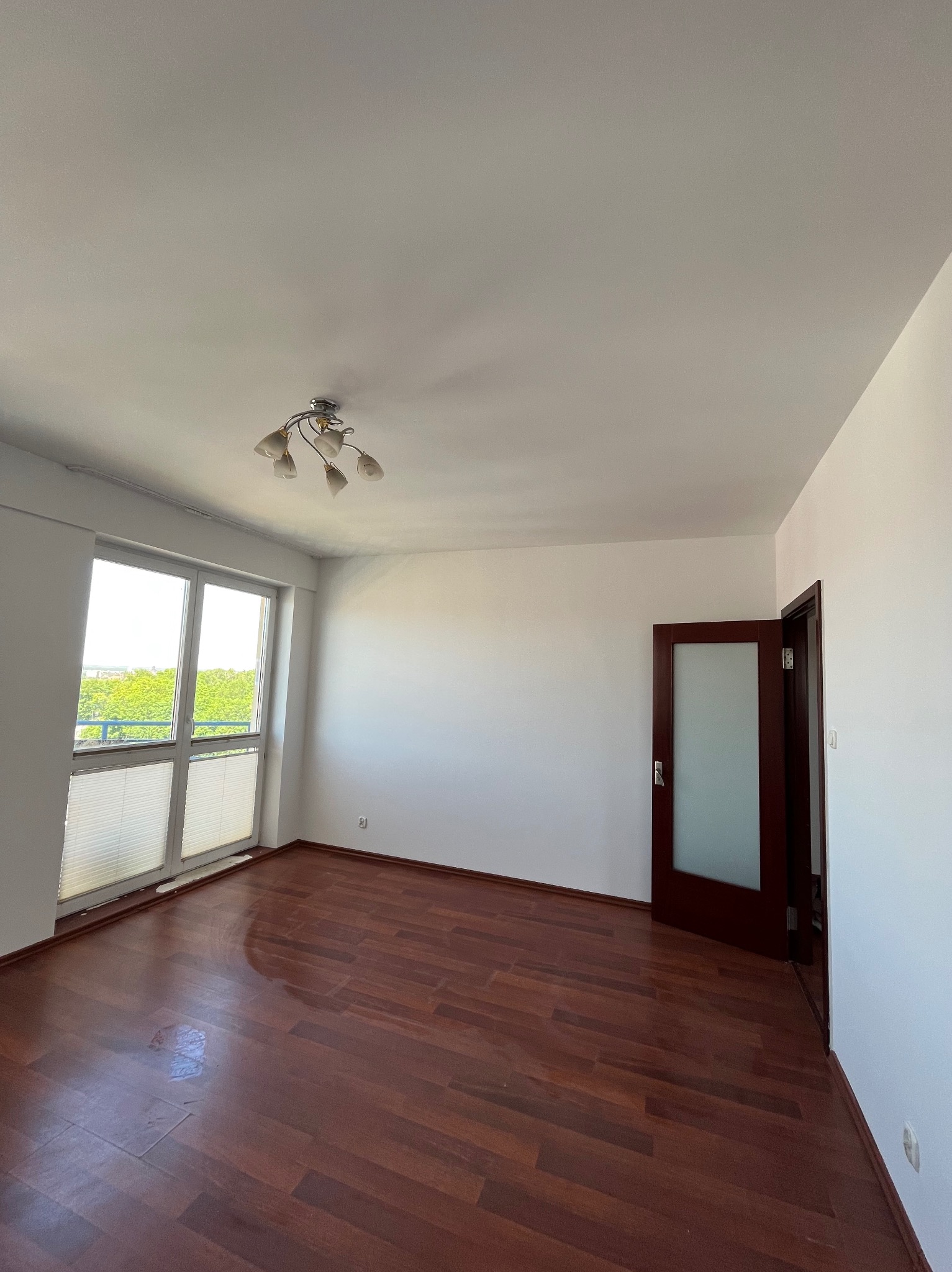 Pusty pokój z drewnianą podłogą, białymi ścianami i oknem z żaluzjami. Widoczny otwarty, ciemny dąb drzwi. Lampa sufitowa w stylu retro. Jasne, naturalne światło.