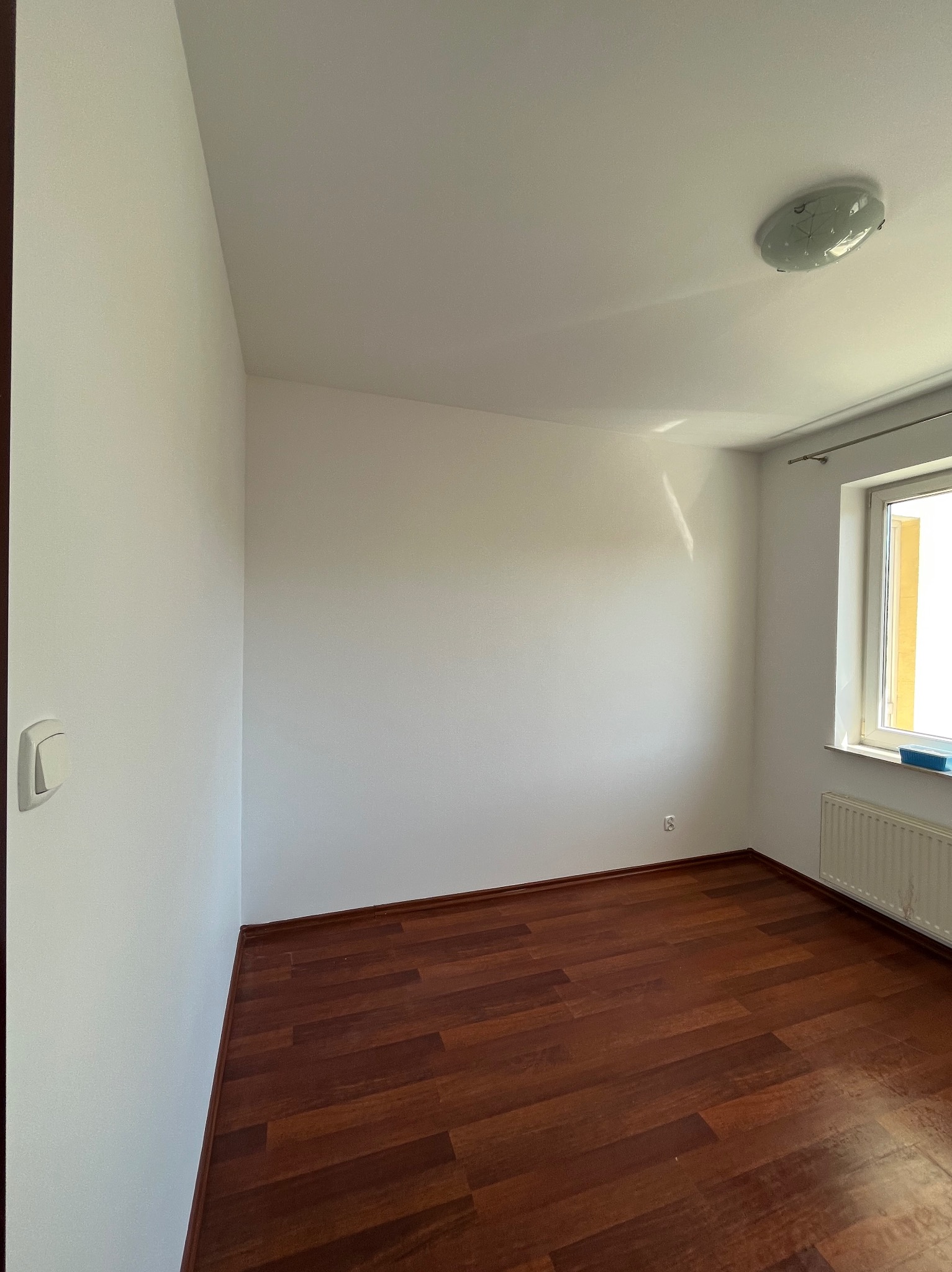 Jasne, puste pomieszczenie po remoncie z brązową podłogą, białymi ścianami i oknem z grzejnikiem. Widoczny włącznik światła i lampa sufitowa.