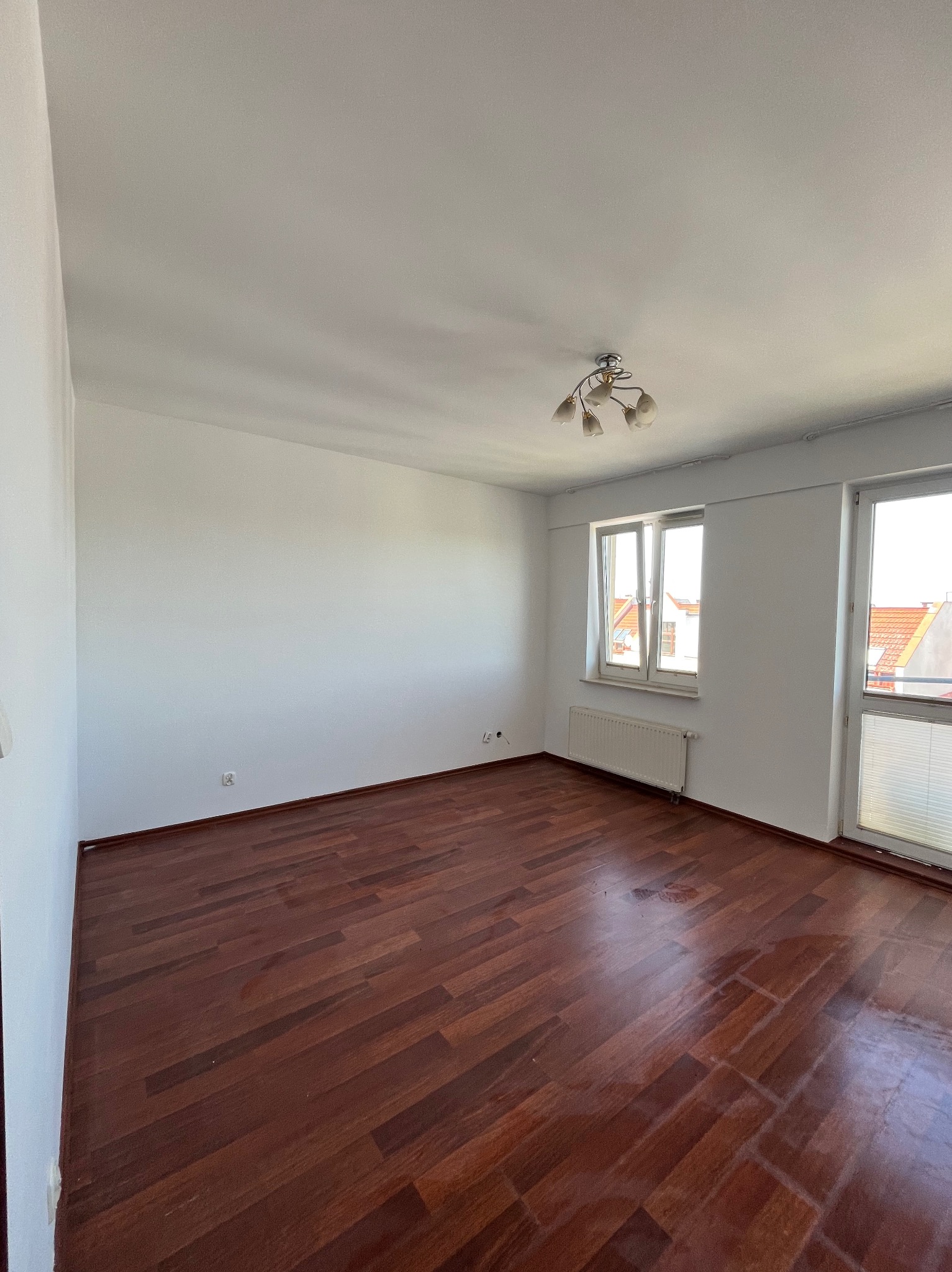 Pusty pokój z parkietem w odcieniach brązu, świeżo pomalowanymi na biało ścianami i oknami z widokiem na zewnątrz. Na suficie widoczna lampa.