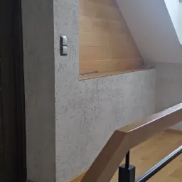Wykończone poddasze z oknem dachowym, ściany pokryte dekoracyjnym tynkiem strukturalnym w odcieniach szarości, drewniana boazeria w skosie dachu oraz fragment balustrady schodowej z drewnianym...