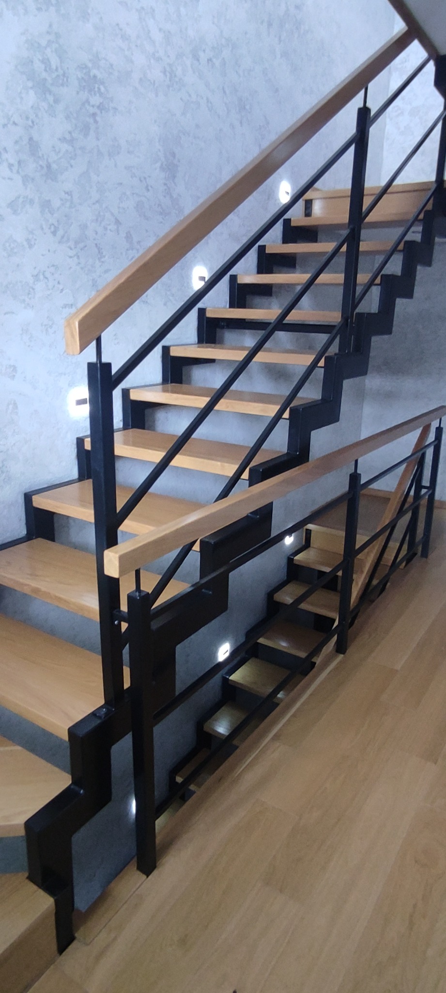 Nowoczesne schody z drewnianymi stopniami i czarną metalową balustradą, oświetlone punktowymi lampkami w ścianie o fakturze betonu.