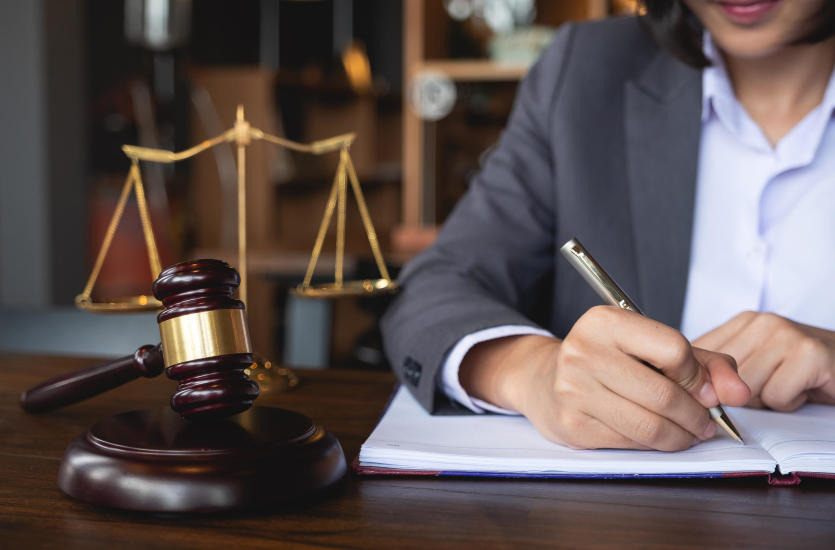 Osoba w szarym garniturze pisze srebrnym długopisem w otwartym notatniku na drewnianym biurku. W tle widoczne złote wagi sprawiedliwości i drewniany młotek sędziowski.