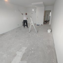 Dagmara Gumna - Mężczyzna maluje ścianę na biało za pomocą wałka na długim uchwycie, stojąc na drabinie w pustym, jasnym pokoju w trakcie remontu, z widocznymi śladami farby na podłodze.