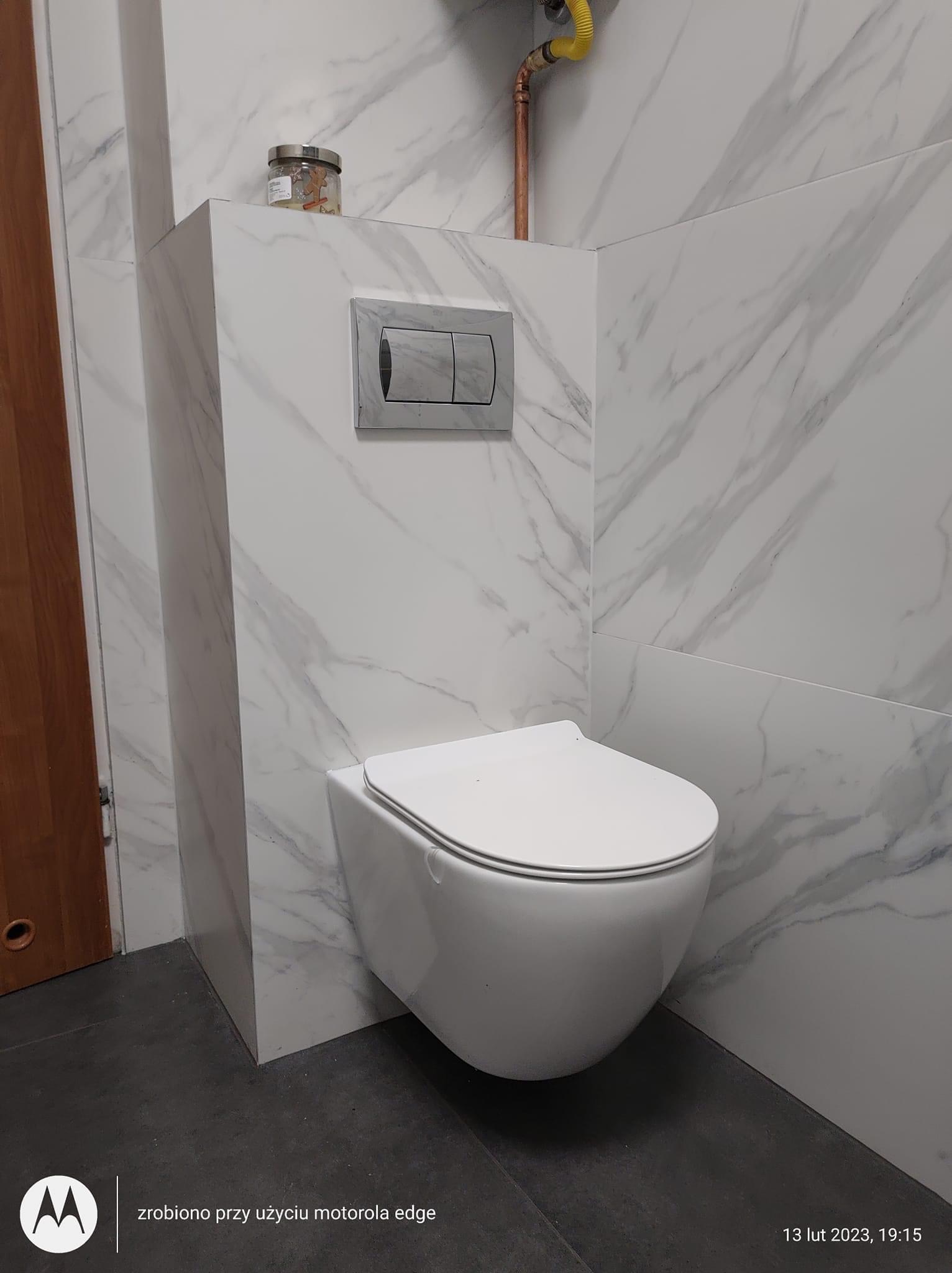 Nowoczesna, podwieszana toaleta z ukrytym spłukiwaniem w zabudowie z płyt imitujących marmur, na tle ściany wykończonej tym samym materiałem. Widoczny fragment instalacji gazowej. Podłoga wyłożona...