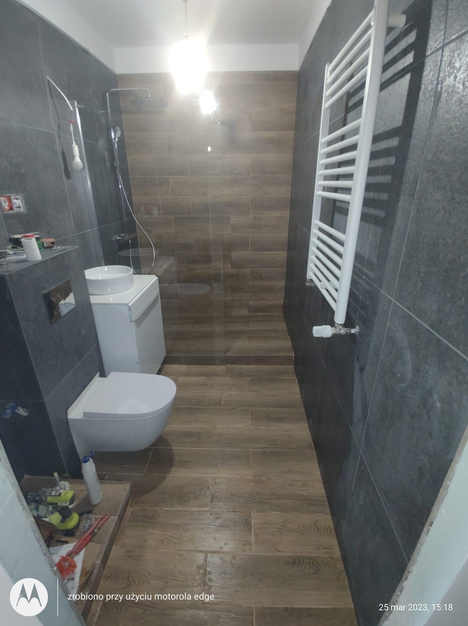 Wnętrze łazienki w trakcie prac wykończeniowych: widoczna toaleta, umywalka nablatowa na szafce, grzejnik drabinkowy, płytki imitujące drewno na podłodze i szare płytki na ścianach, narzędzia...