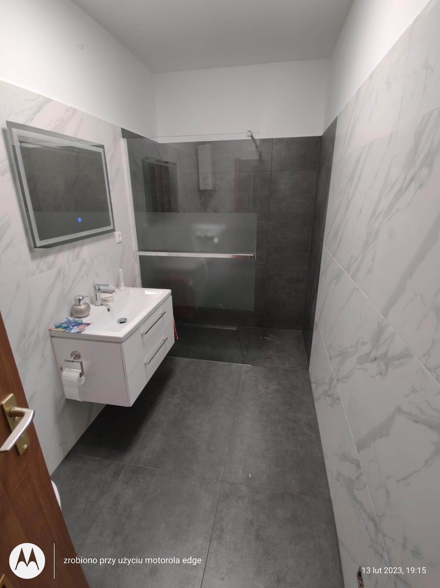 Nowoczesna łazienka z umywalką nablatową, podświetlanym lustrem i kabiną prysznicową z szarymi płytkami, wykończona białymi płytkami imitującymi marmur.