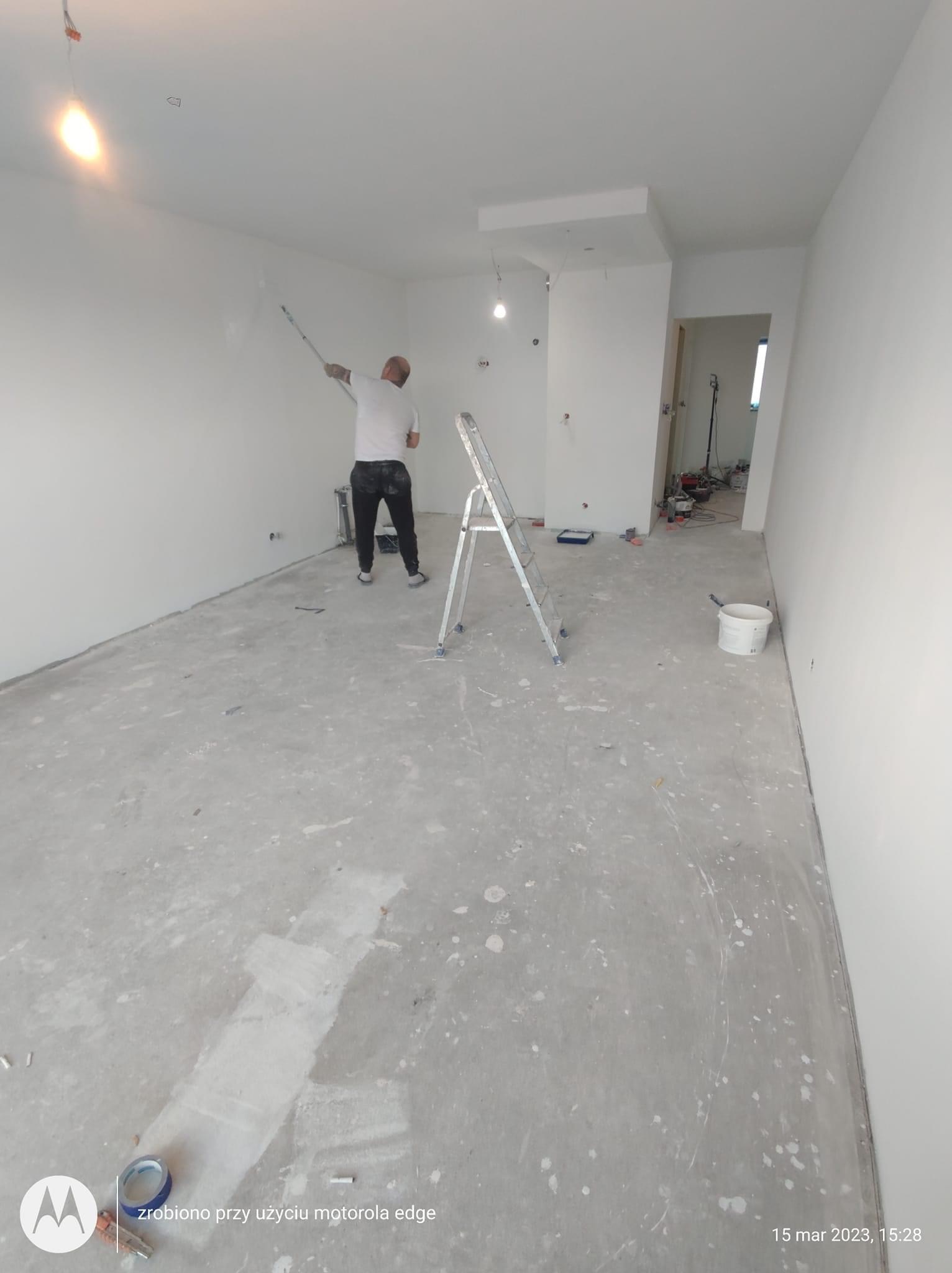 Mężczyzna maluje ścianę na biało za pomocą wałka na długim uchwycie, stojąc na drabinie w pustym, jasnym pokoju w trakcie remontu, z widocznymi śladami farby na podłodze.