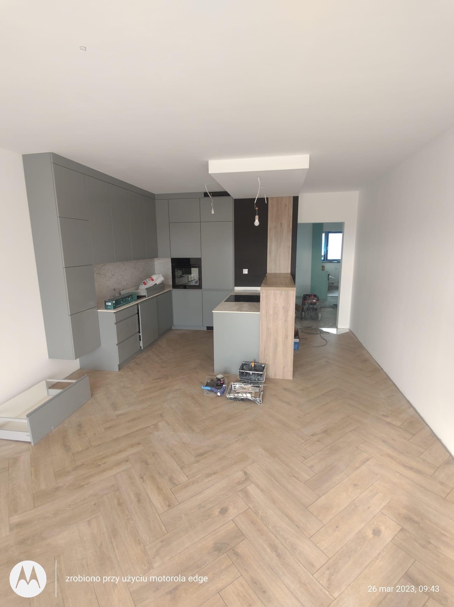 Nowoczesna kuchnia w trakcie montażu, z szarymi szafkami, drewnianym blatem wyspy i parkietem ułożonym w jodełkę. Widoczne narzędzia i pozostałości po pracach.