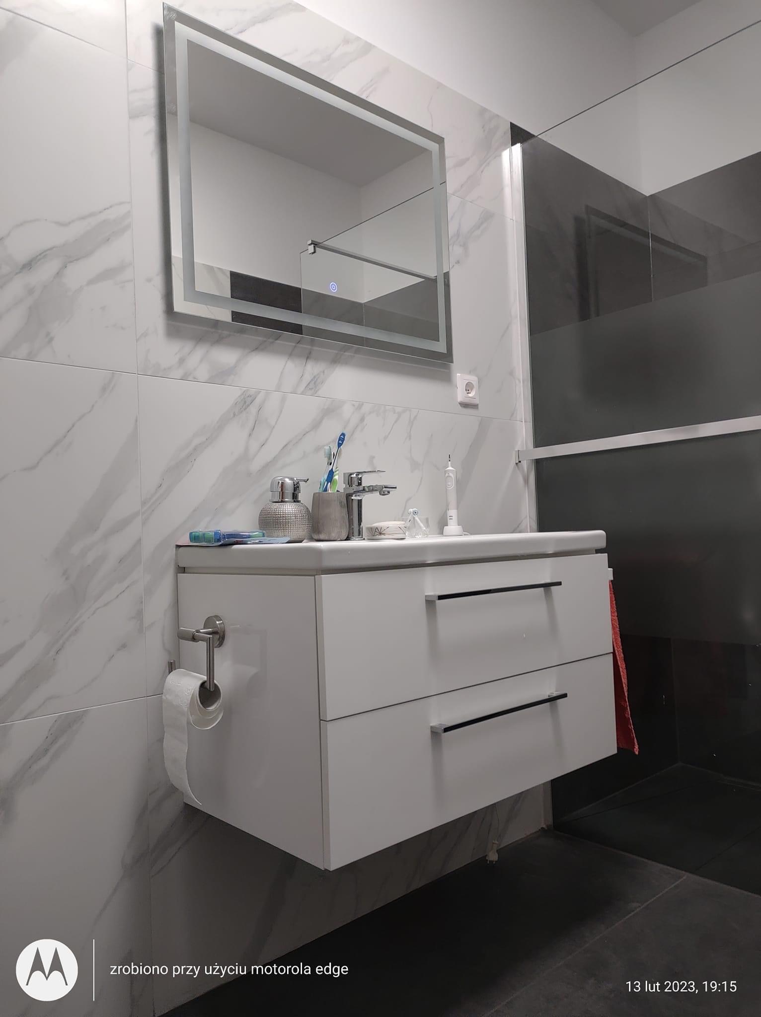 Nowoczesna łazienka z białymi płytkami imitującymi marmur, wisząca szafka z umywalką, podświetlane lustro i kabina prysznicowa z ciemnego szkła.