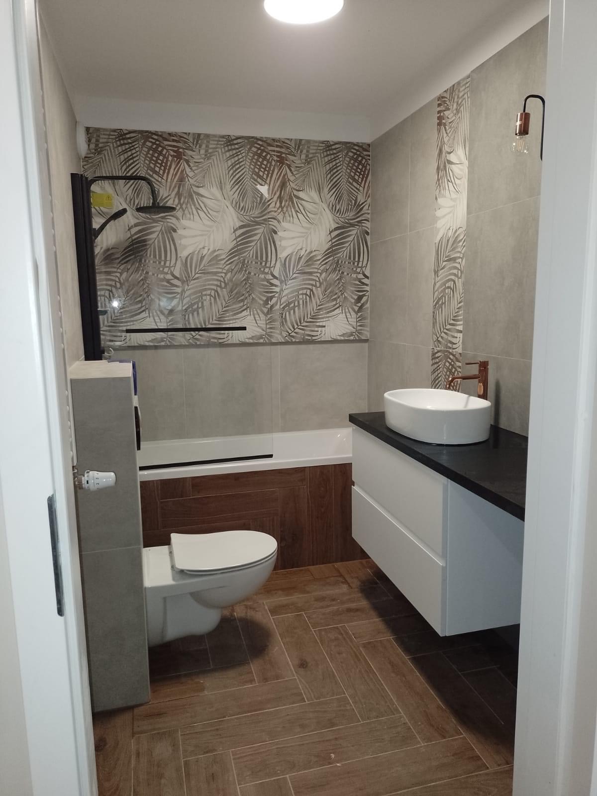 Nowoczesna łazienka z drewnopodobną podłogą w jodełkę, umywalką nablatową na białej szafce z czarnym blatem, toaletą oraz wanną z parawanem, zdobiona tapetą w liście.