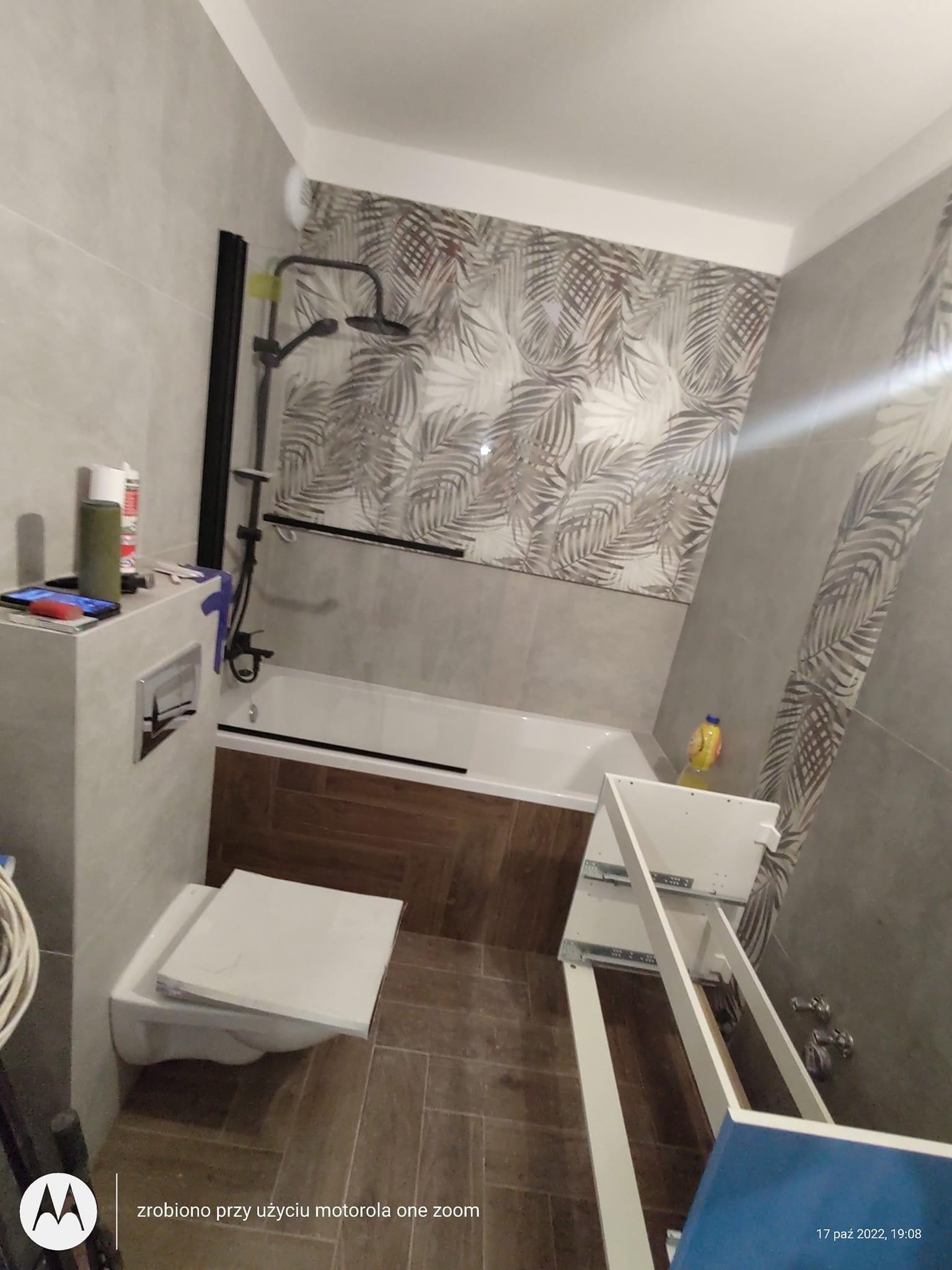 Łazienka w trakcie prac: widoczna wanna z czarną armaturą, częściowo ułożone płytki z motywem liści, podwieszana toaleta z otwartą klapą oraz elementy szafki łazienkowej w dolnym rogu.