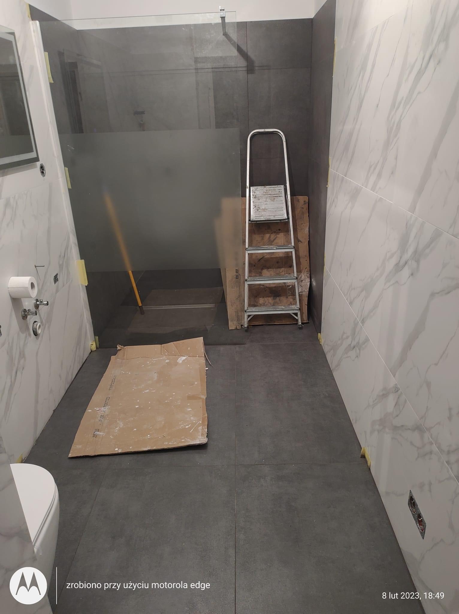Wnętrze łazienki w trakcie remontu: widoczne płytki imitujące marmur na ścianach, szara podłoga, fragment toalety, kabina prysznicowa z częściowo matową szybą, drabina oparta o ścianę...