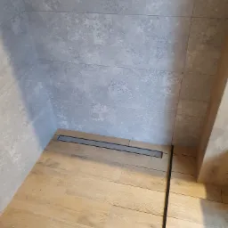 Nowoczesna kabina prysznicowa z odpływem liniowym, wykończona płytkami imitującymi drewno na podłodze i szarymi płytkami na ścianach.