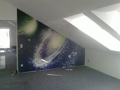 Poddasze z tapetą kosmiczną na ścianie skośnej, dwa okna dachowe w suficie, szara wykładzina na podłodze. Widoczne gniazdka elektryczne na ścianie.