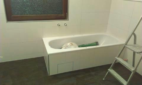 Biała wanna obudowana płytkami w łazience z brązową podłogą i oknem. Aluminiowa drabina oparta o bok wanny. Widoczne akcesoria łazienkowe.