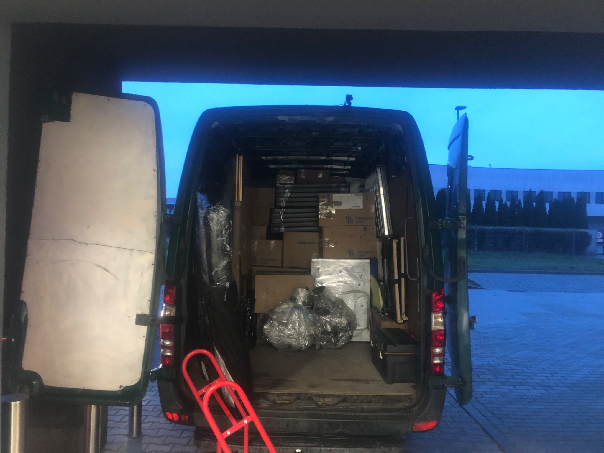 Wnętrze busa załadowane kartonami, meblami i sprzętem AGD, przygotowane do transportu, z widocznym czerwonym wózkiem transportowym z tyłu pojazdu.