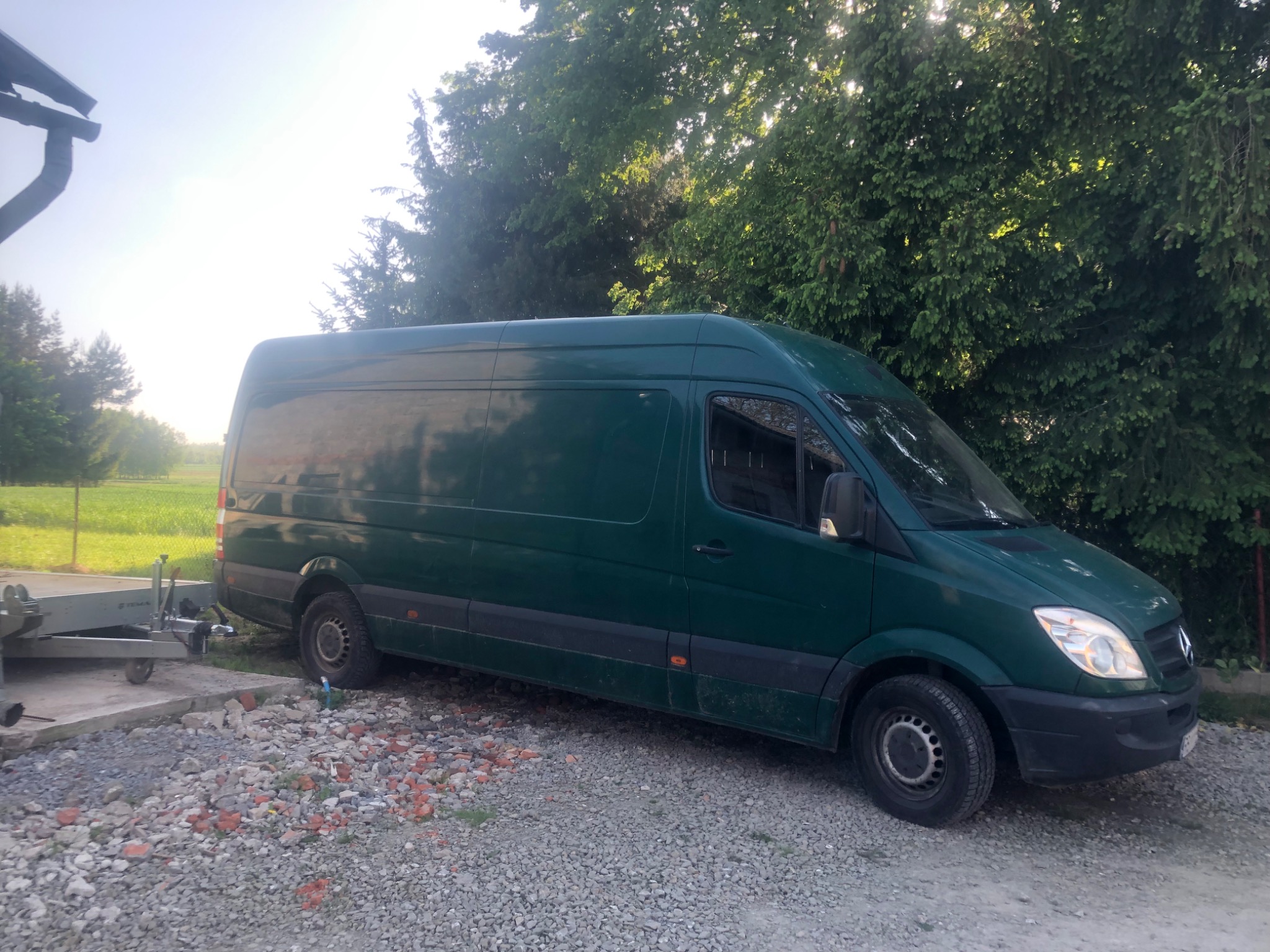 Ciemnozielony bus Mercedes-Benz Sprinter z doczepioną przyczepką, zaparkowany na żwirowym podłożu z cegłami, na tle drzew i pola.