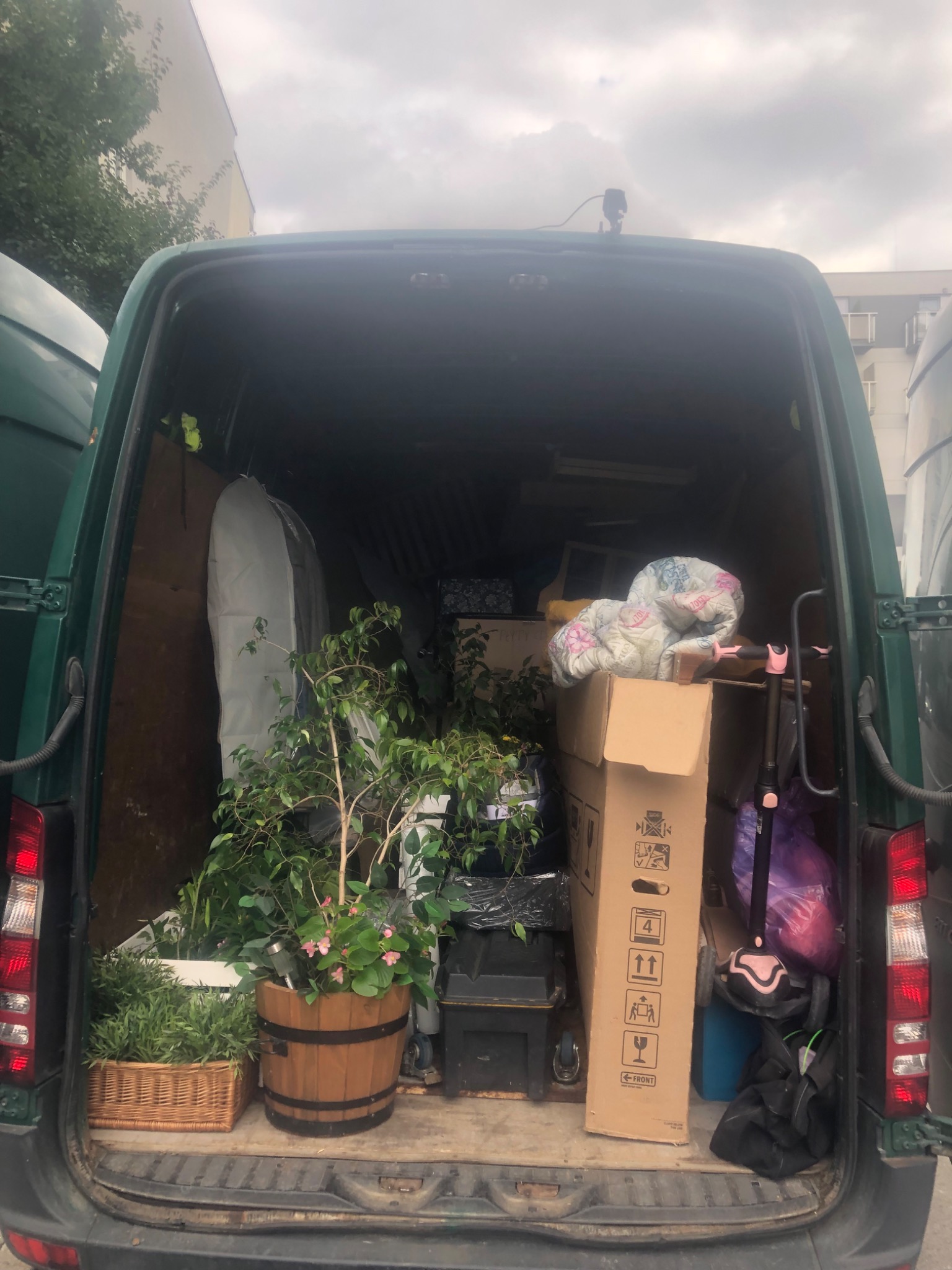 Wnętrze zielonego vana załadowane roślinami doniczkowymi, kartonami i innymi przedmiotami gotowymi do transportu. Widoczny duży drewniany kwietnik z różowymi kwiatami.