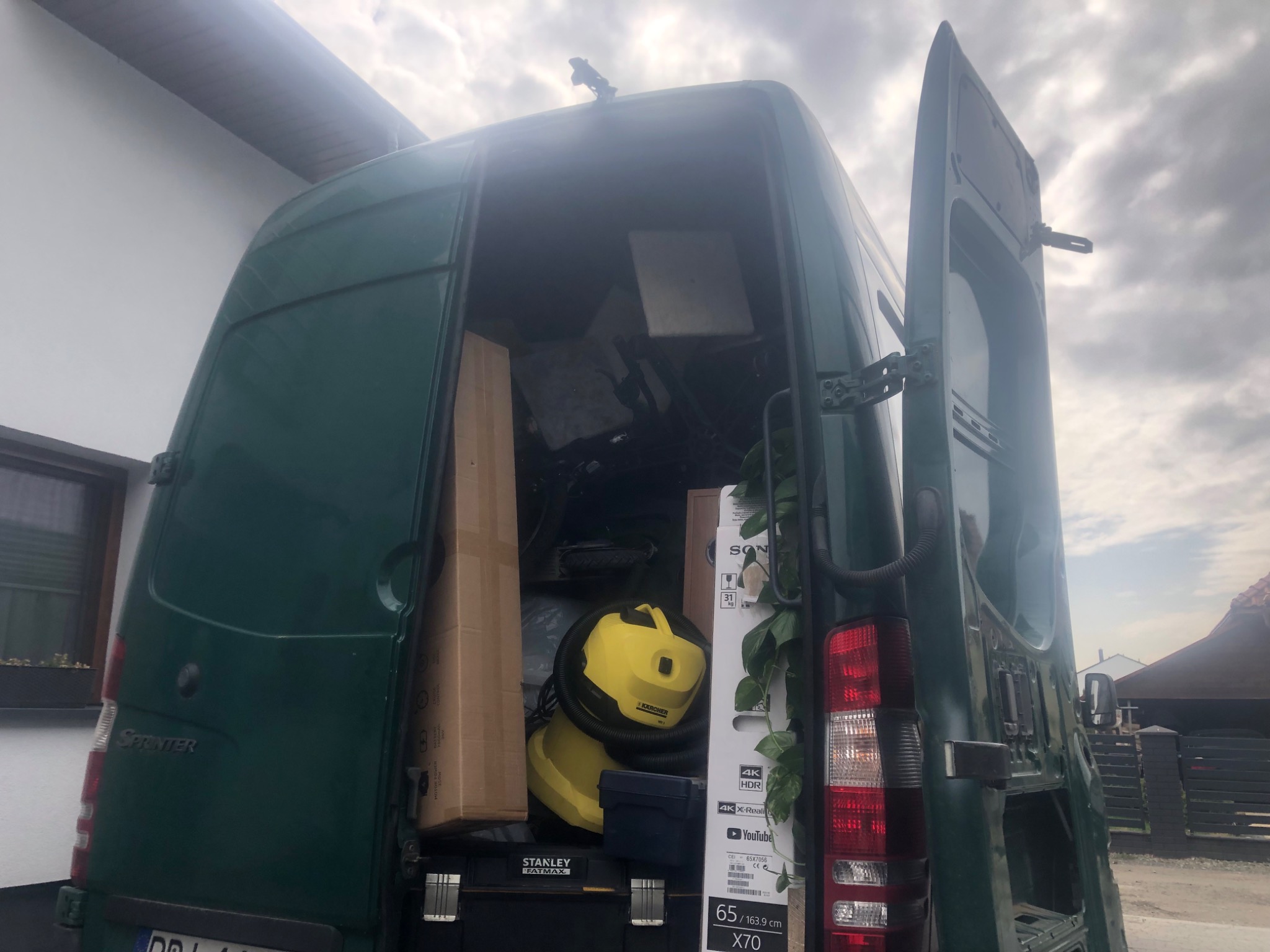 Otwarty bagażnik zielonego samochodu dostawczego marki Sprinter, wypełniony kartonami, odkurzaczem Karcher, skrzynką narzędziową Stanley Fatmax i telewizorem Sony 4K X70 przygotowany do transportu.