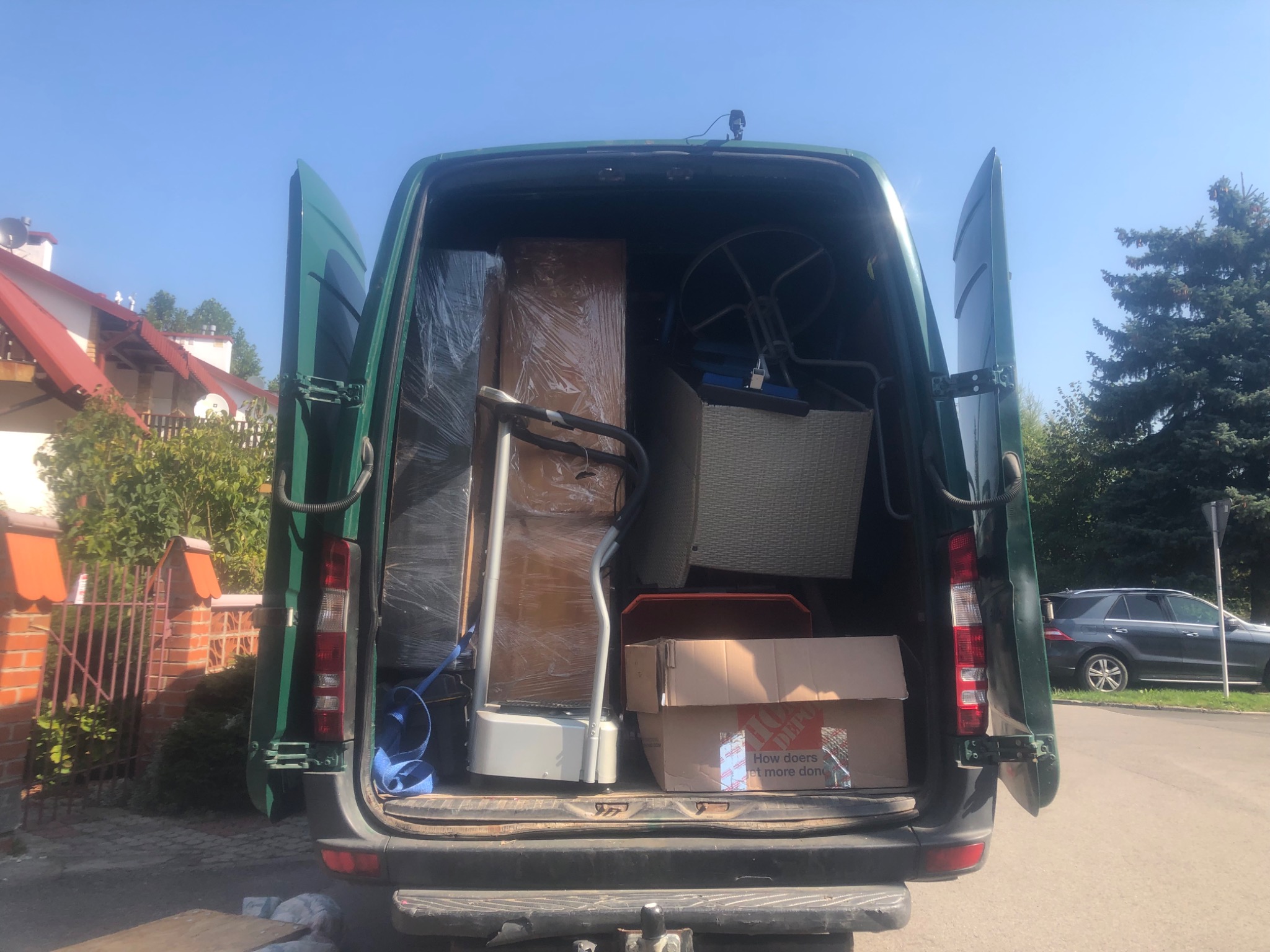 Wnętrze zielonego busa z otwartymi drzwiami, załadowane meblami i sprzętami, w tym bieżnią i zapakowanymi kartonami, widok na dom z czerwonym dachem i zaparkowany samochód osobowy.