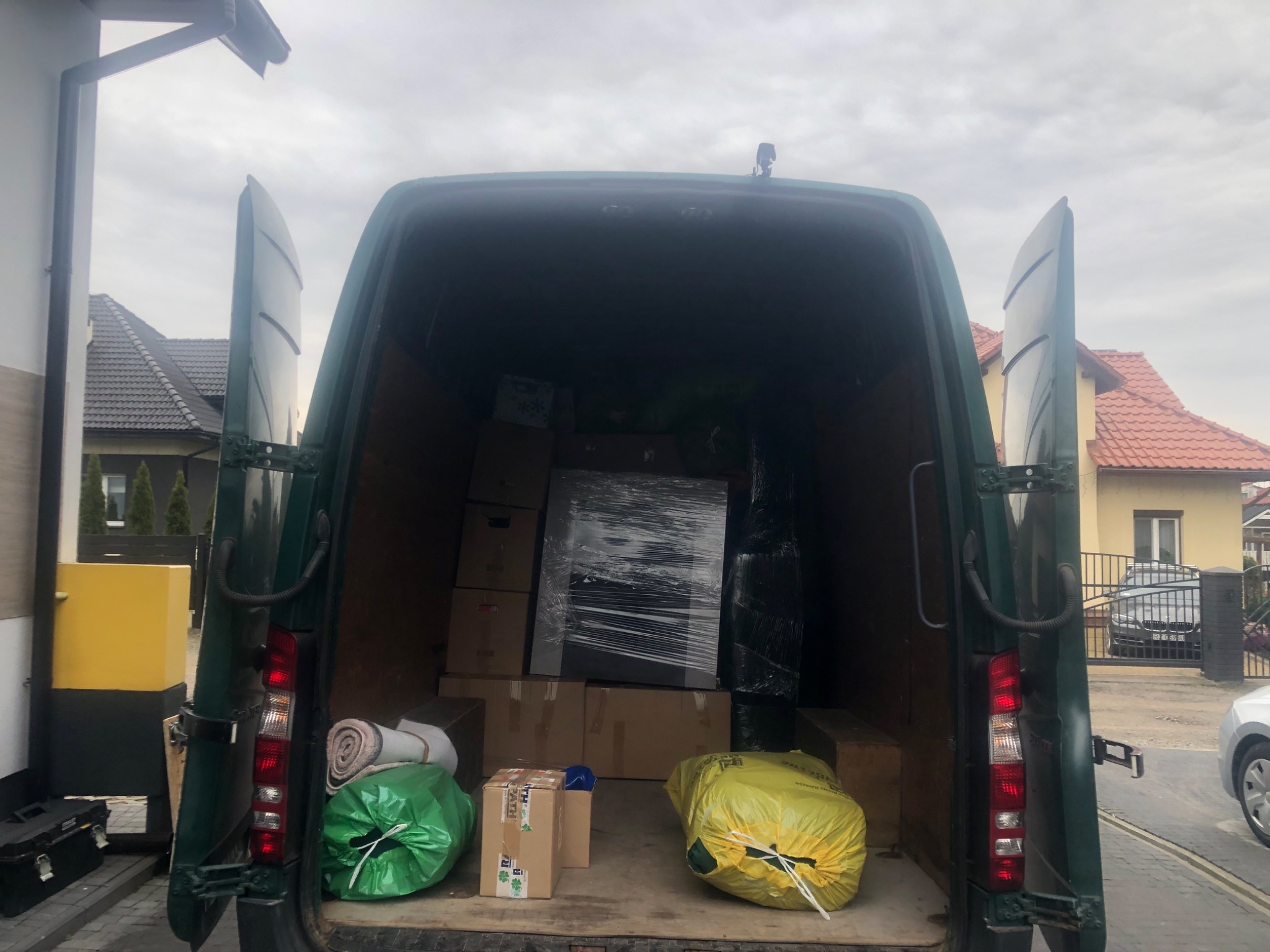 Otwarty tył zielonego samochodu dostawczego wypełnionego kartonami i meblami owiniętymi folią stretch, widok z zewnątrz na domy jednorodzinne w tle.
