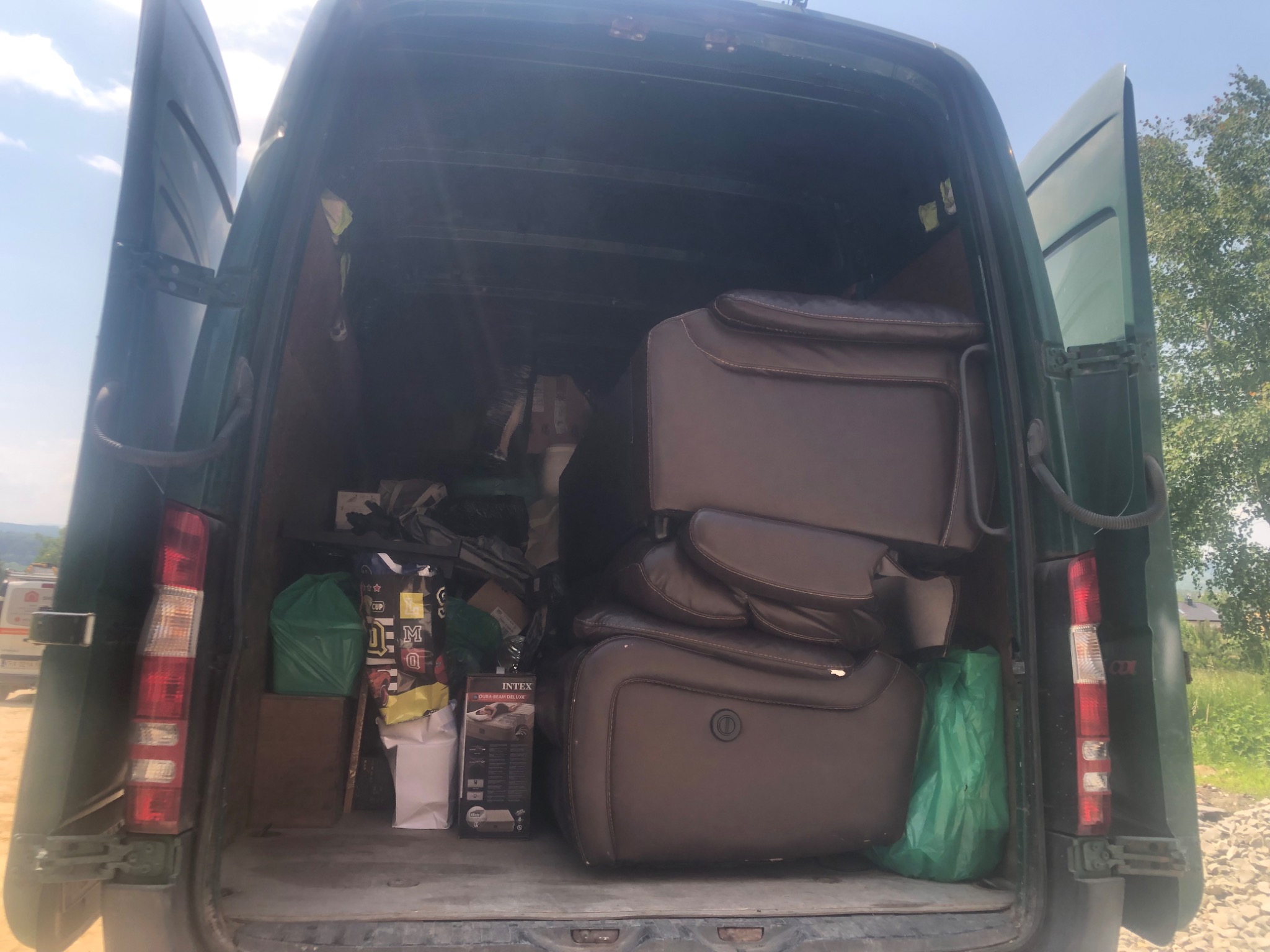 Wnętrze otwartego busa wypełnione brązowymi fotelami masującymi, widoczne pudełka i worki z zakupami, w tle zielona trawa i drzewa.