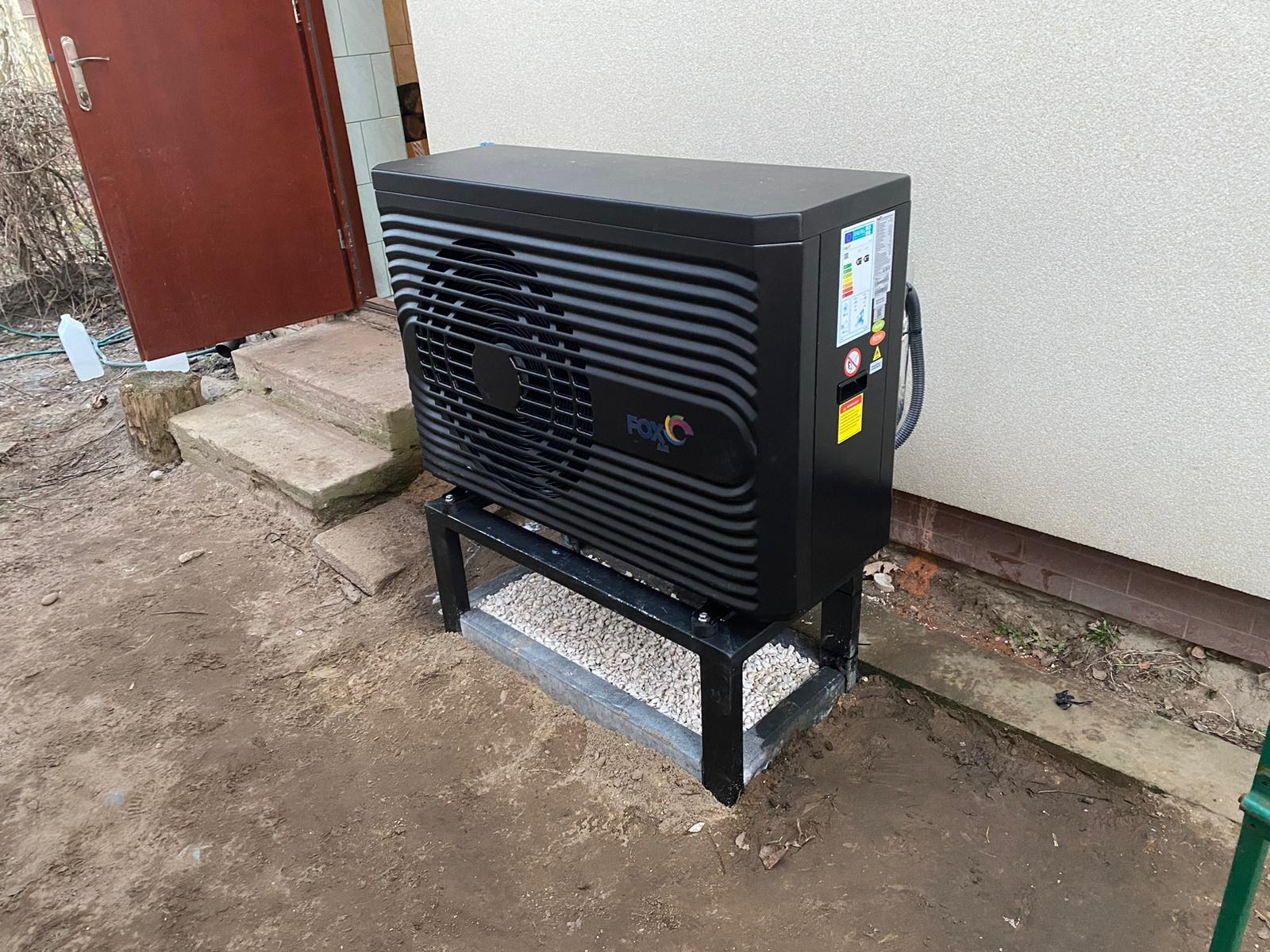 Czarna pompa ciepła marki FOX na metalowym stelażu, umieszczona na zewnątrz budynku, z widocznymi etykietami energetycznymi i osłoną wentylatora.
