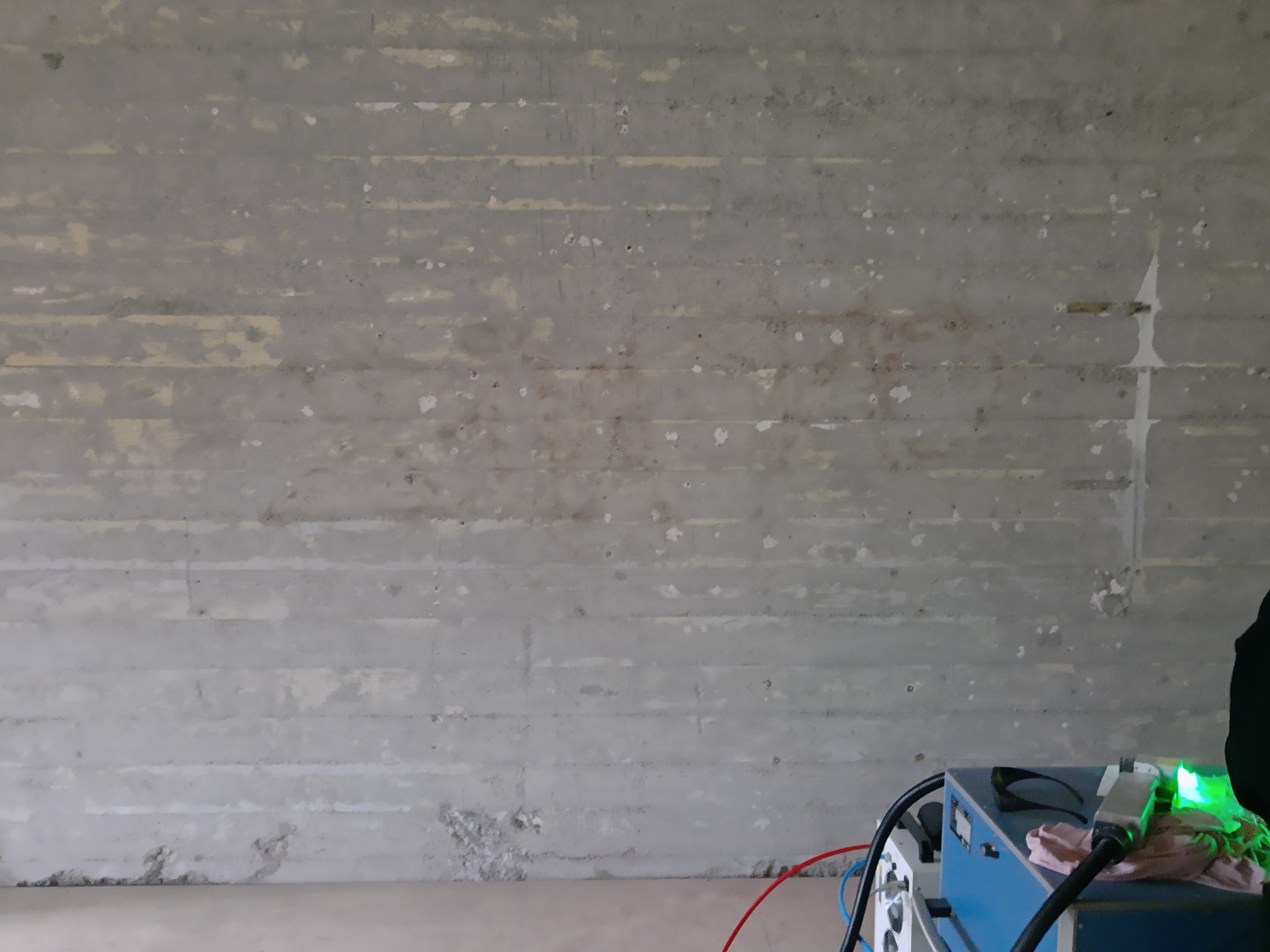 Szara betonowa ściana z poziomymi śladami po deskowaniu, częściowo oczyszczona, z widocznymi plamami i przebarwieniami, w dolnej części kadru niebieska maszyna z przewodami i zielonym światłem.