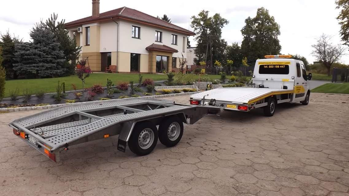 Laweta z dwuosiową przyczepą na tle domu jednorodzinnego z zadbanym ogrodem, widok z tyłu.