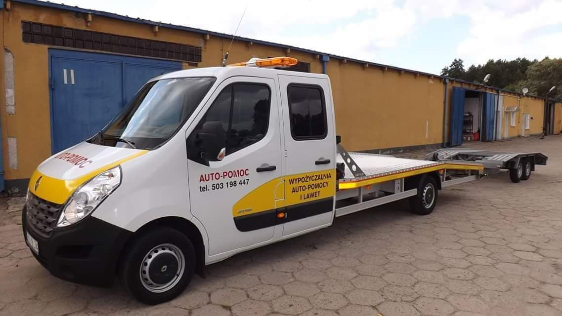 Biała laweta Renault Master z żółtymi akcentami i napisem 'AUTO-POMOC tel. 503 198 447, WYPOŻYCZALNIA AUTO-POMOCY I LAWET' zaparkowana na betonowym placu przed żółtym budynkiem z niebieskimi bramami.