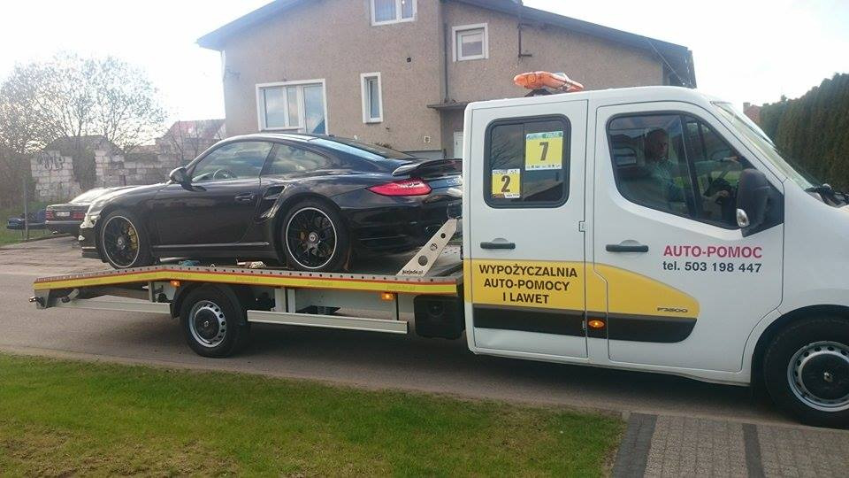 Czarny sportowy samochód Porsche na lawecie auto-pomocy z żółtym pasem i napisem 'WYPOŻYCZALNIA AUTO-POMOCY I LAWET' na tle budynku mieszkalnego.
