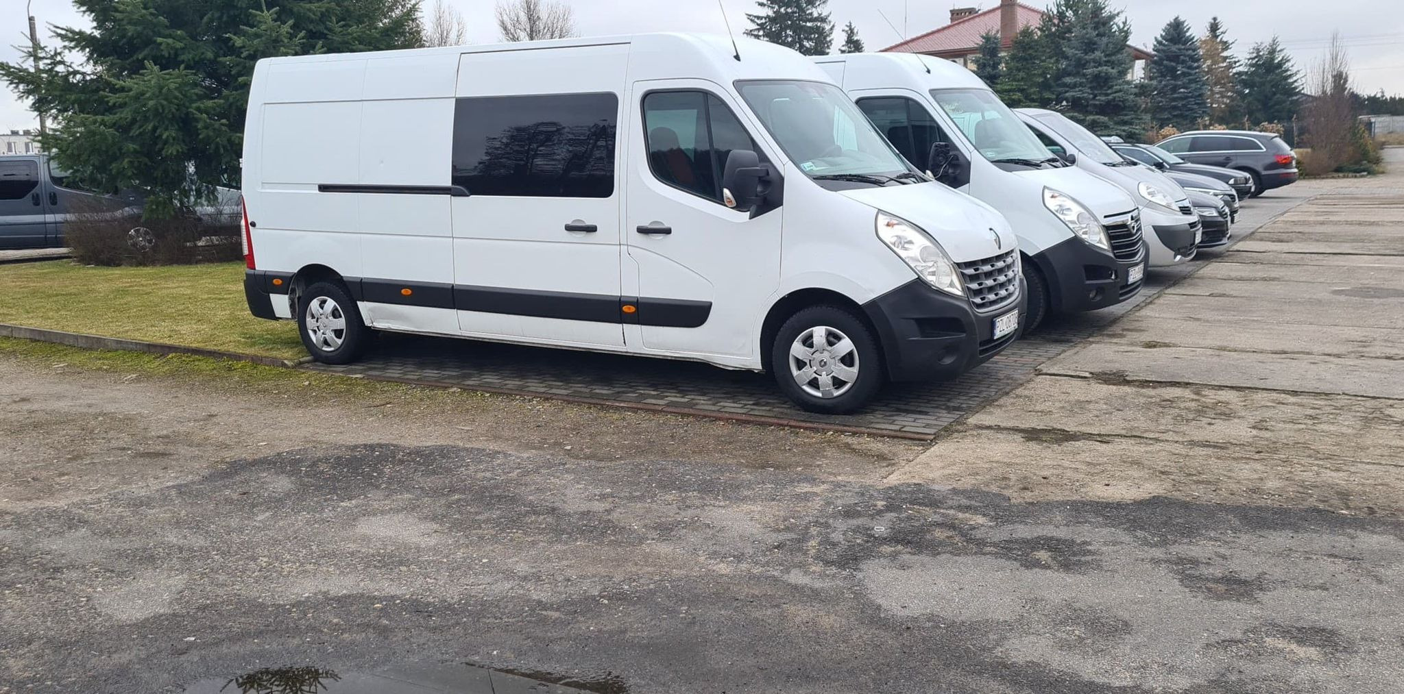 Rząd białych samochodów dostawczych Renault Master zaparkowanych na zewnątrz na betonowym placu, widok z boku na pierwszy pojazd z czarnymi szybami.
